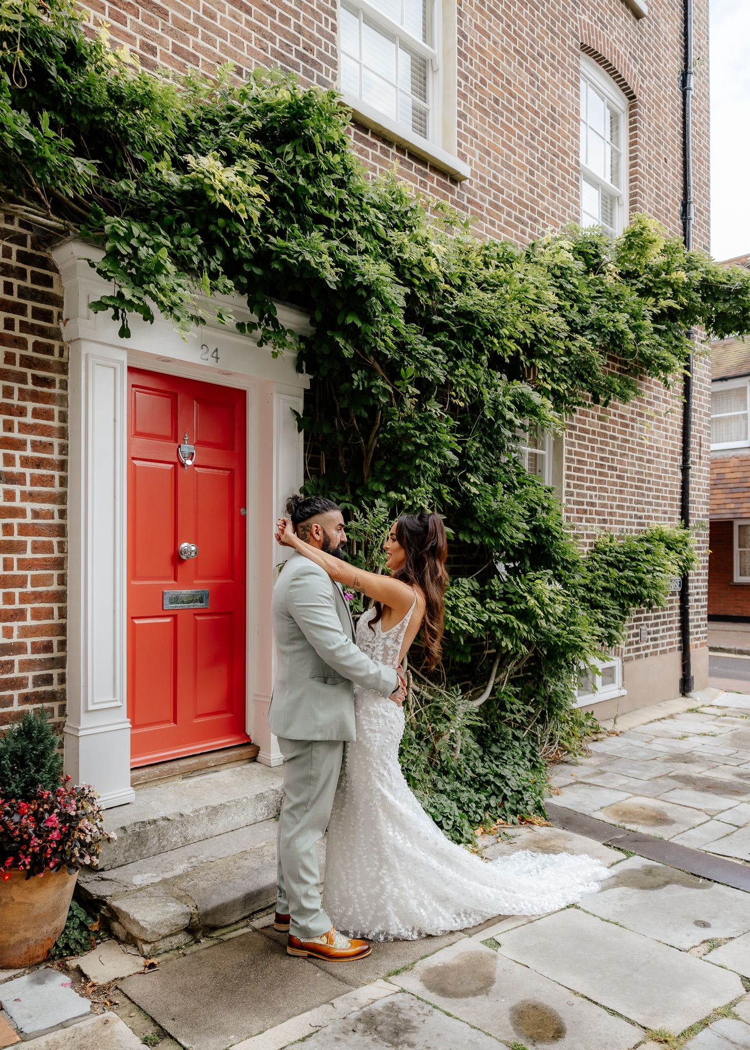 A couple in wedding attire dances and poses in front of a brick building with red doors and climbing ivy.
