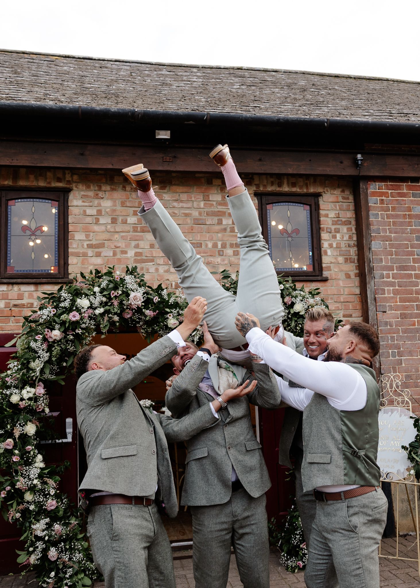 Wedding party in gray suits celebrating with energetic jumping poses against brick wall backdrop.