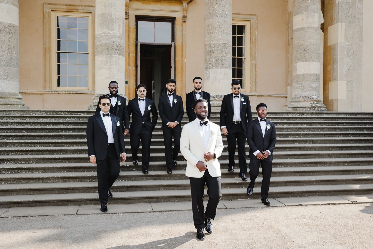 Wedding party members in formal black tuxedos and white gowns walk down grand stone steps of an elegant building.