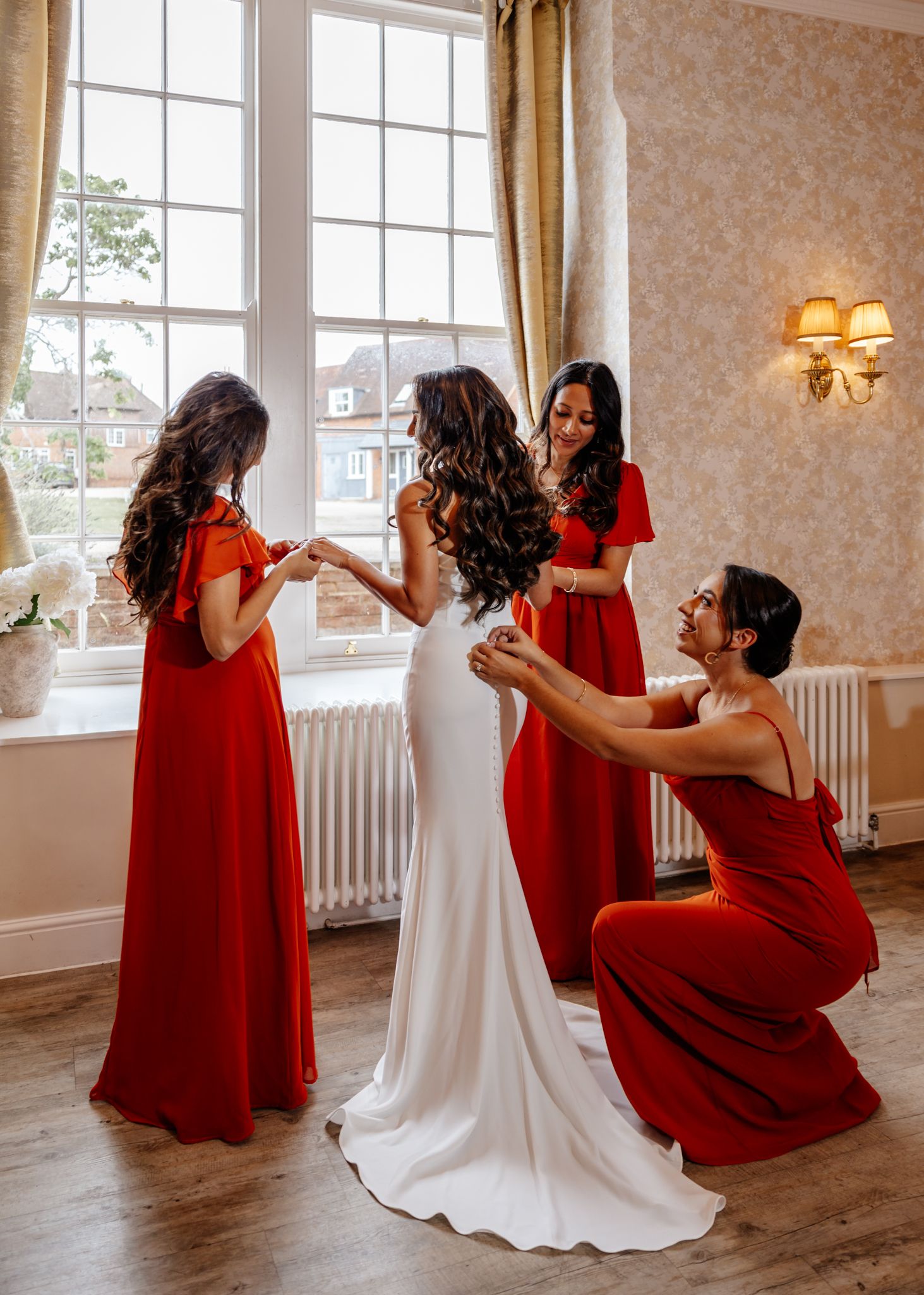 A bridal party in red floor-length dresses helps adjust a white wedding gown by a bright window in an elegant room.