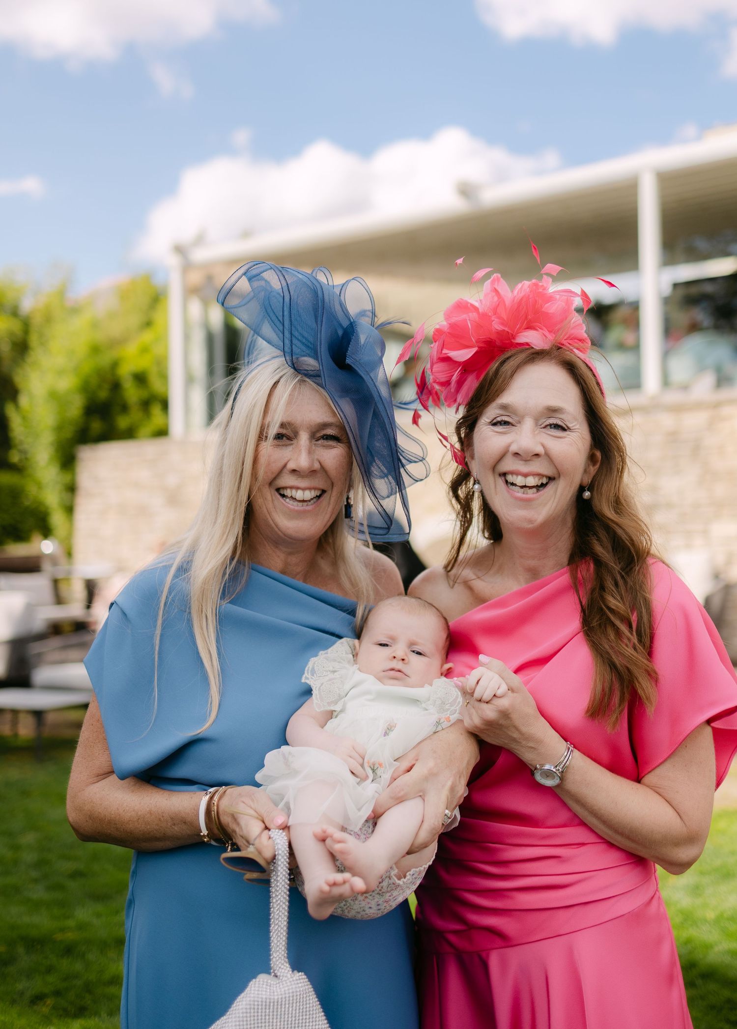 Two women in colorful fascinators and dresses smiling outdoors while holding a baby at a garden event.