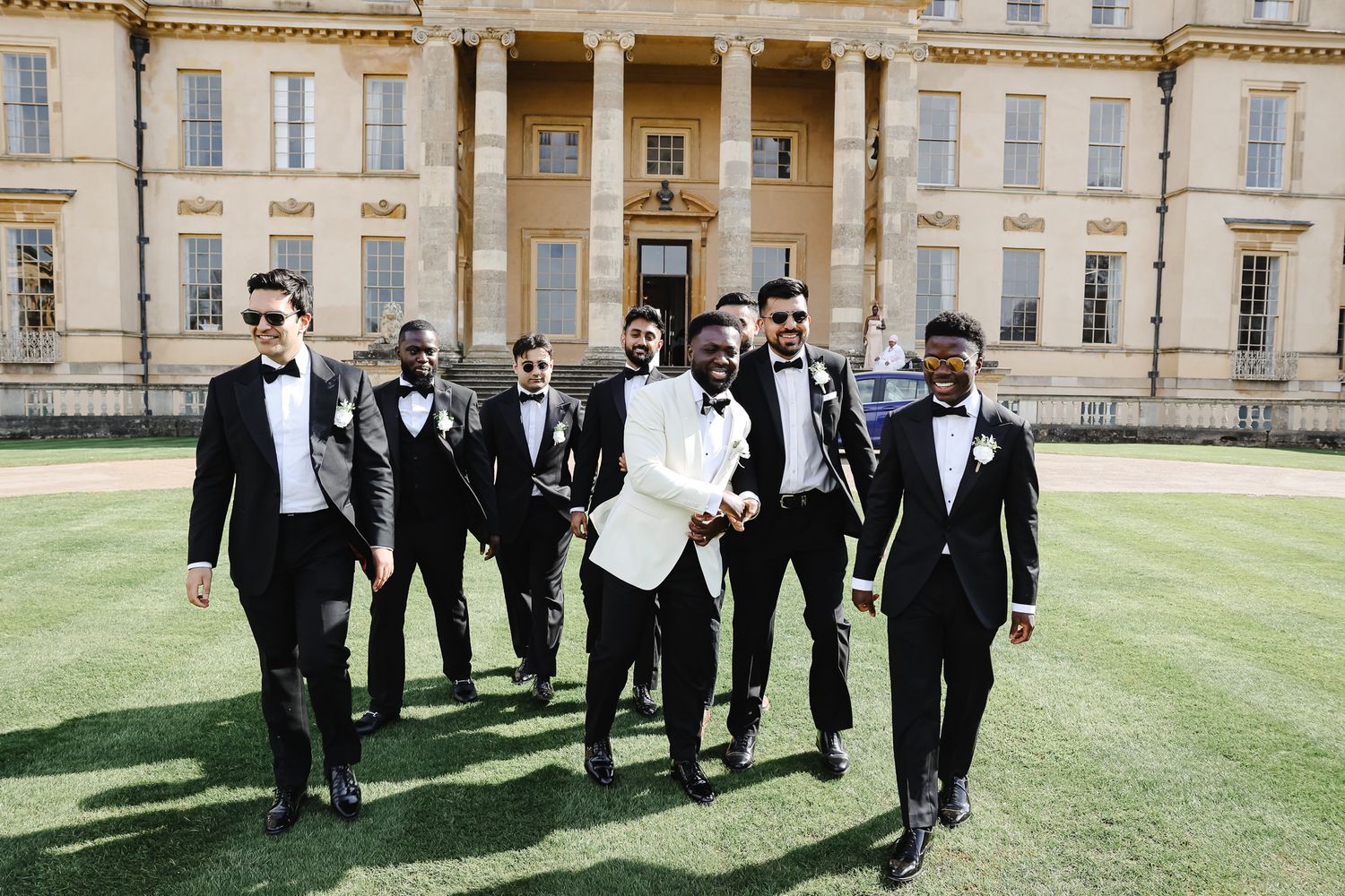 Groomsmen in formal attire walk and pose playfully on manicured grounds of elegant venue.