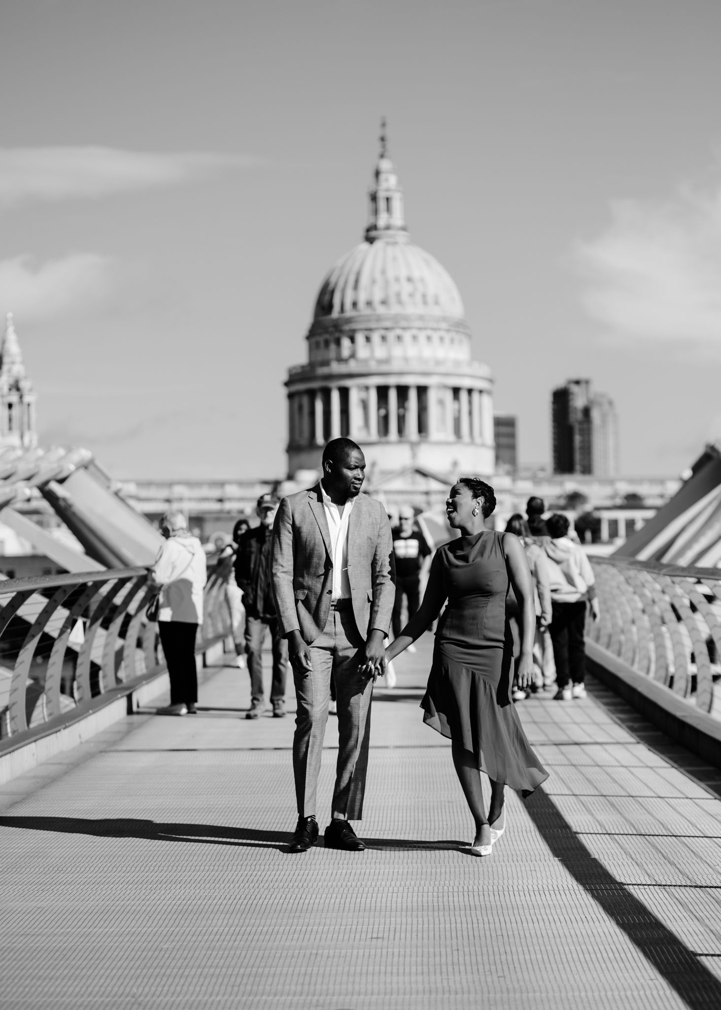 Black and white photo of a couple holding hands on Millennium Bridge with St. Paul's Cathedral in London backdrop.