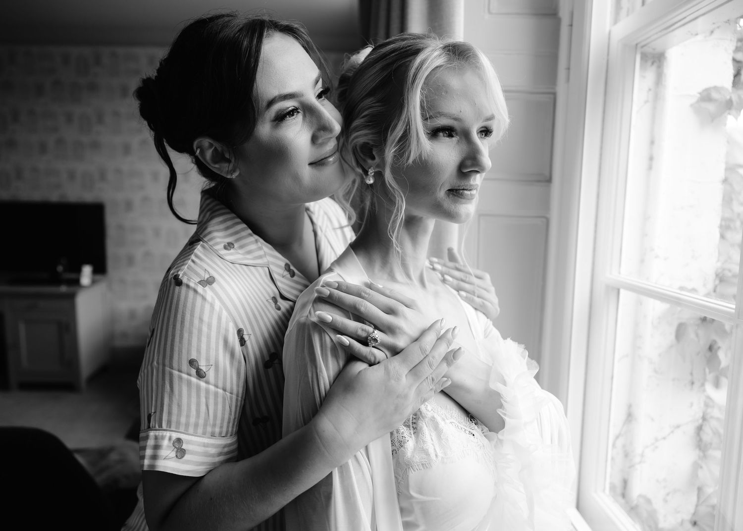 Black and white photo of a bridesmaid helping a bride with her dress near a sunlit window.
