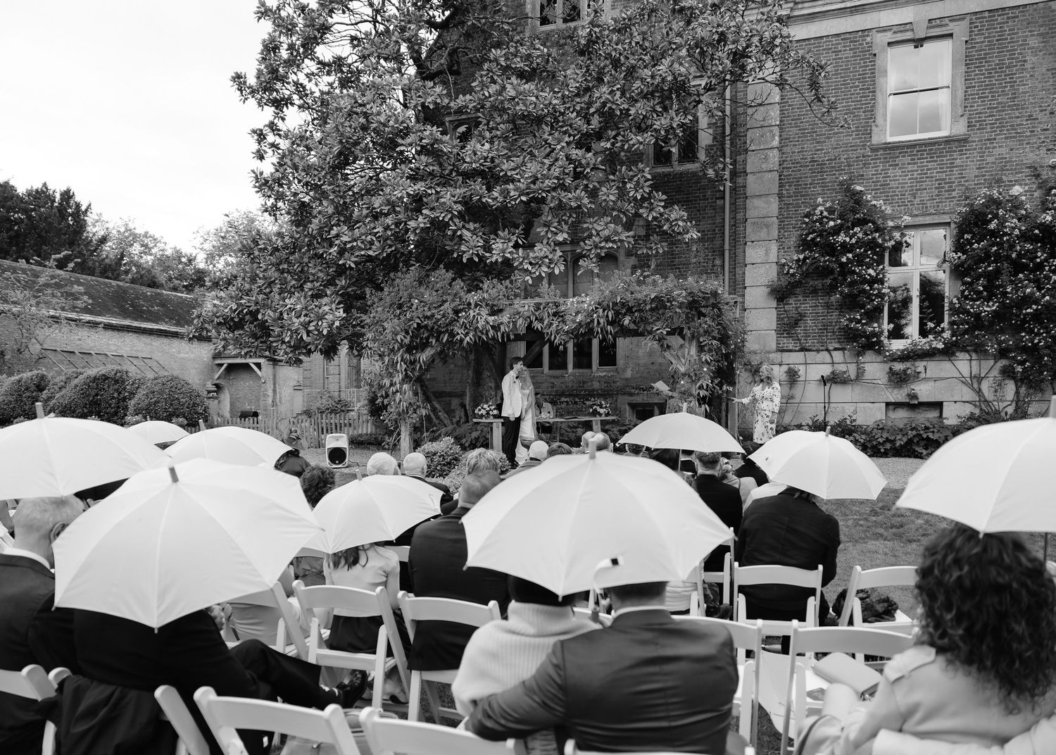Outdoor wedding ceremony in the rain with guests holding white umbrellas seated in white chairs before a historic manor.