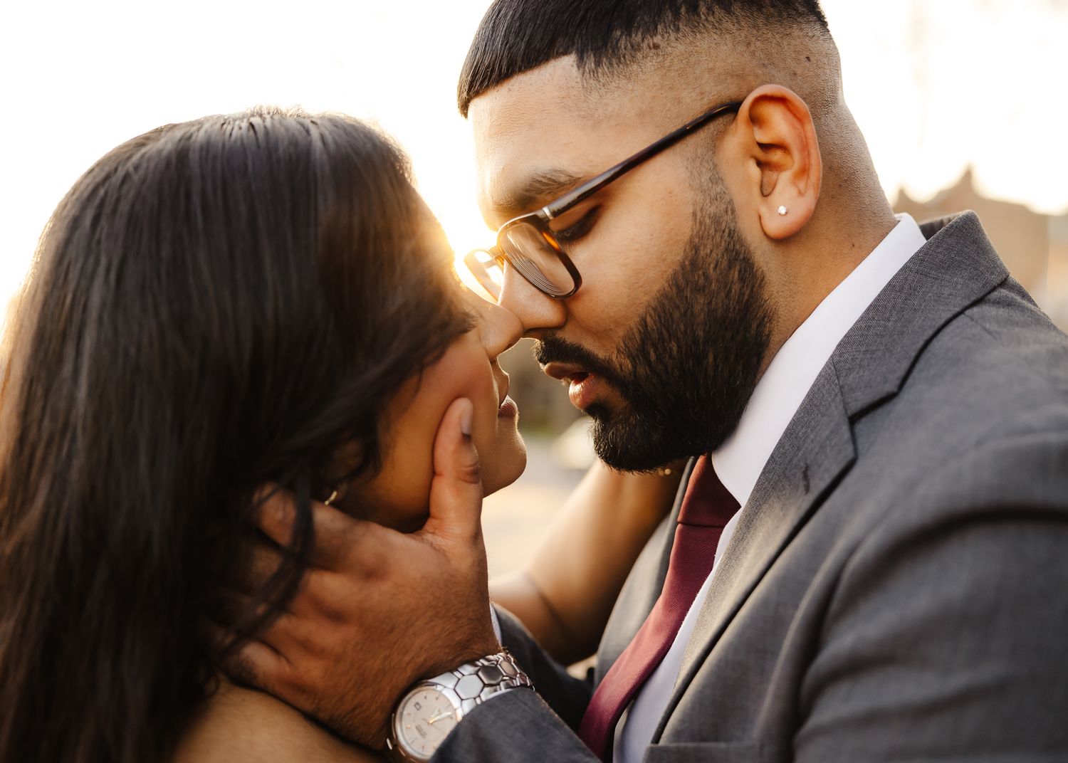 A couple shares a romantic kiss, the man in a gray suit with glasses, backlit by warm golden sunlight.