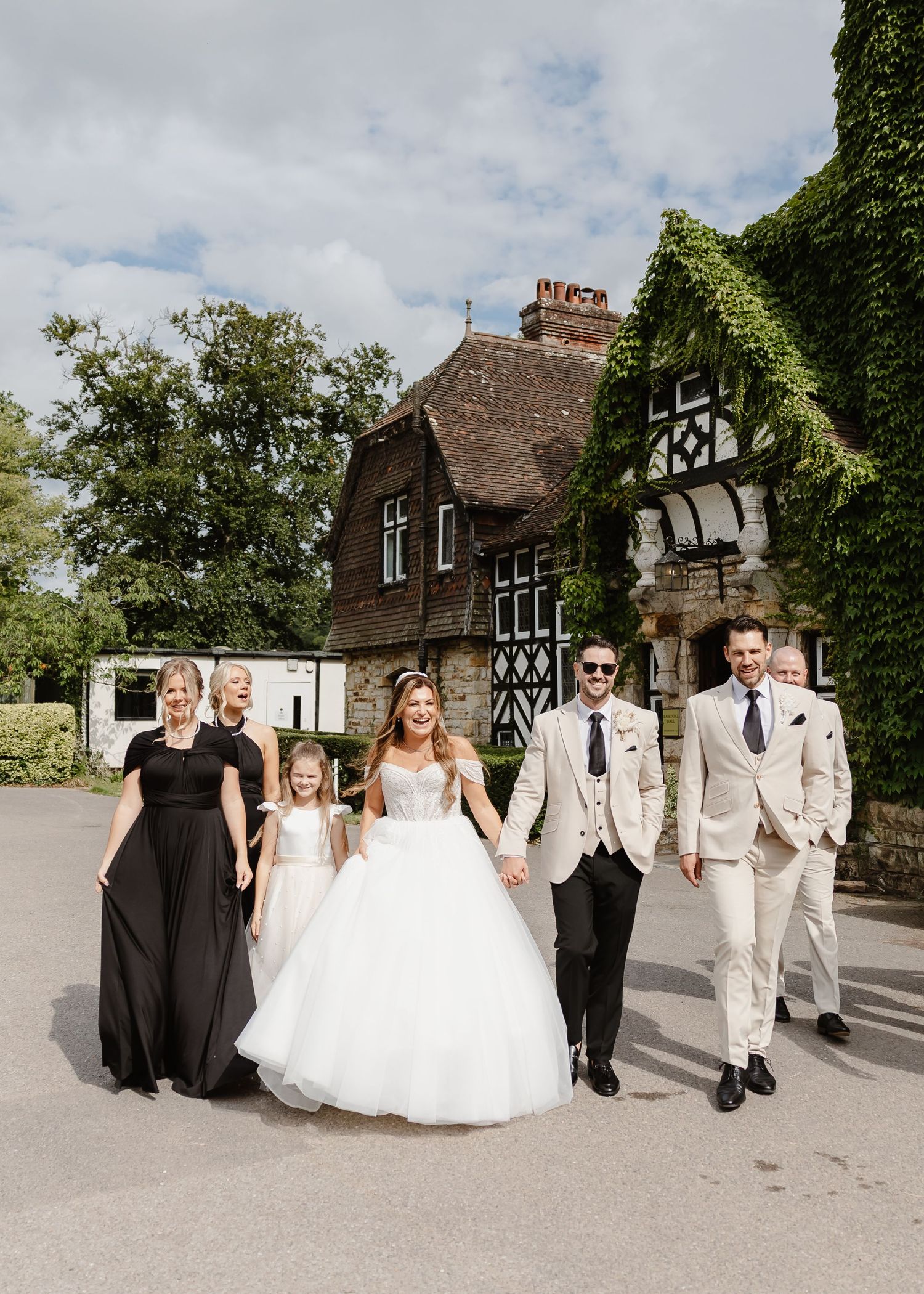 Wedding party walks outdoors near ivy-covered Tudor building, bride in white gown, bridesmaids in black, groomsmen in beige suits.