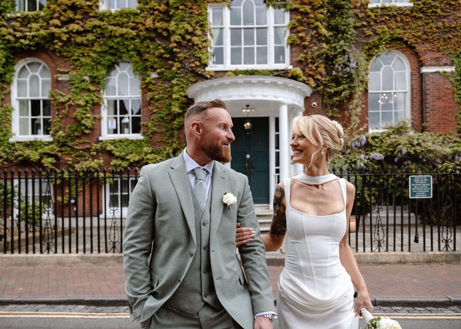 Bride and groom in elegant wedding attire standing outside an ivy-covered historic building on their wedding day.