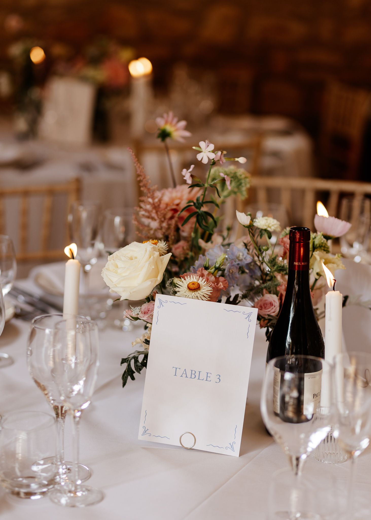 Elegant wedding reception table setting with wine glasses, floral centerpieces, and numbered table cards in soft lighting.