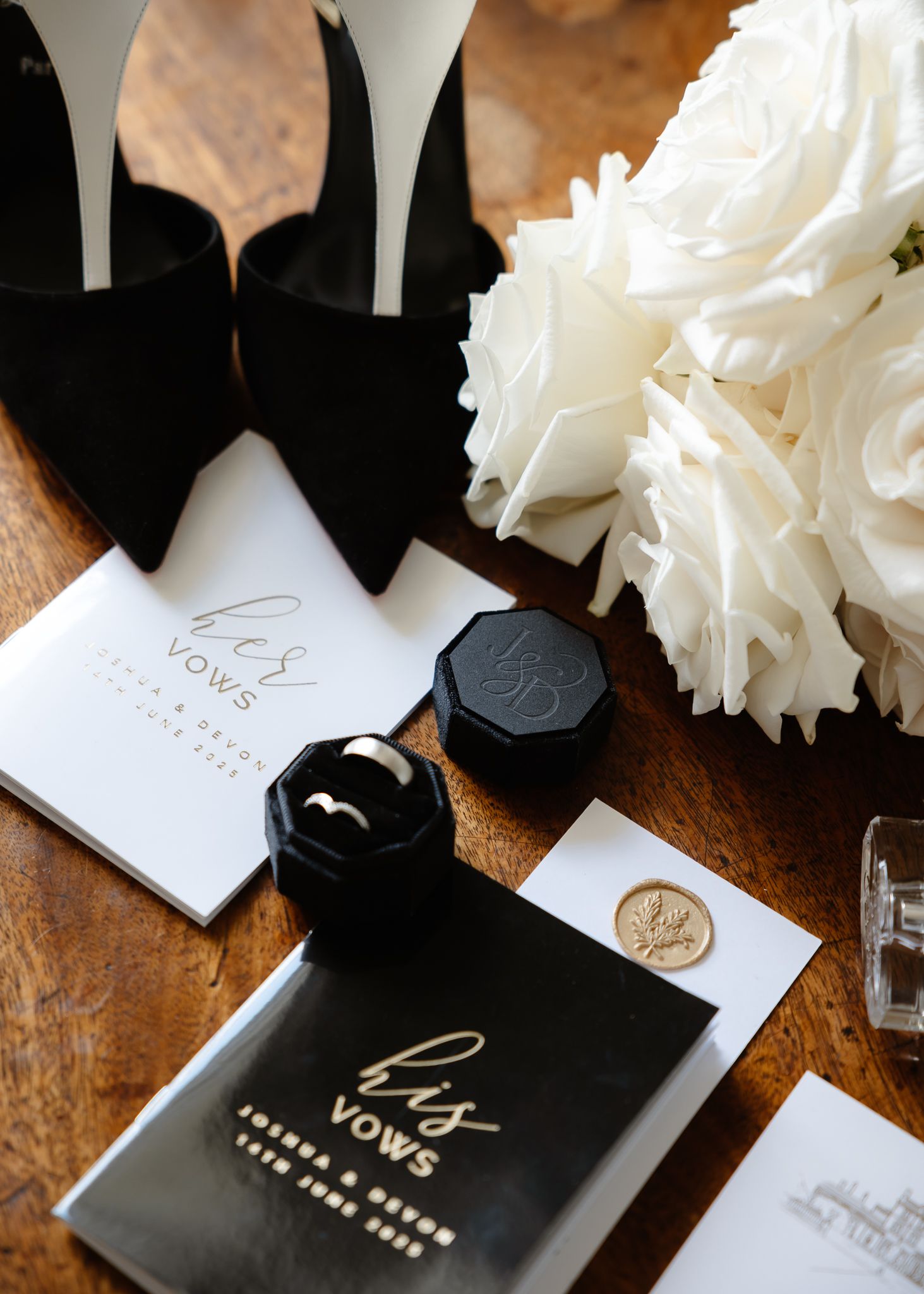 Black and white wedding details including shoes, invitations, wax seals, and white roses on a wooden surface.
