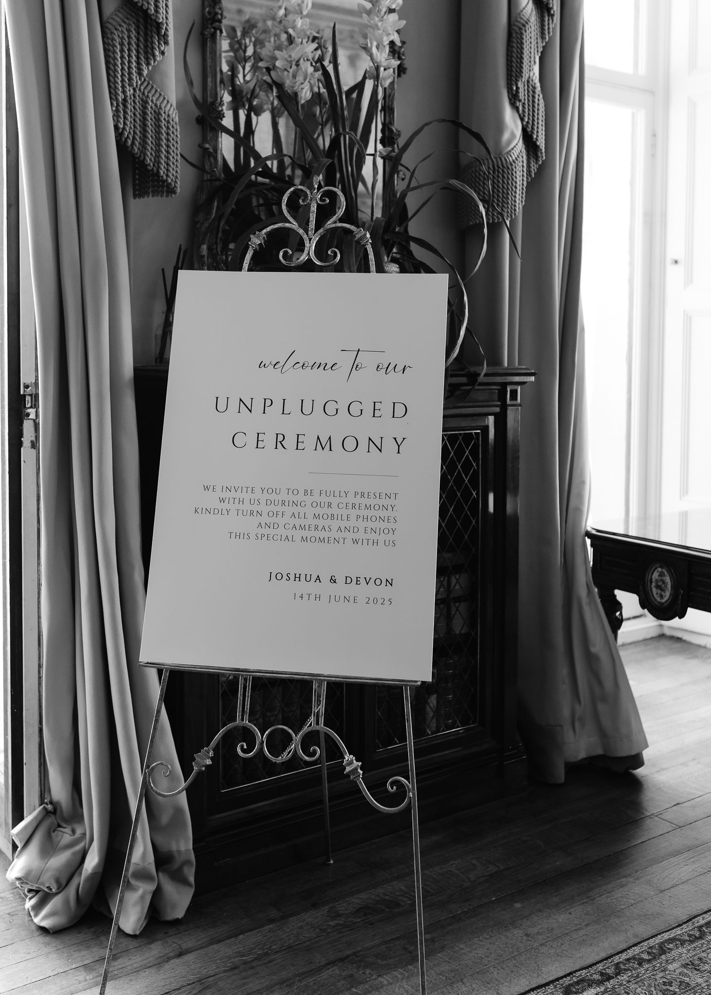 A black and white welcome sign for an unplugged ceremony displayed on an ornate metal easel with curtains in background.
