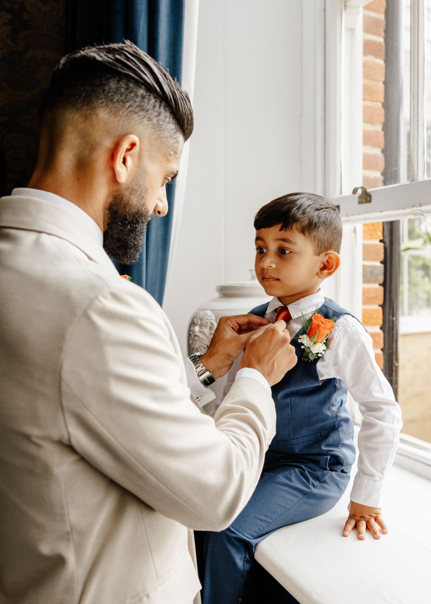 A person in a cream blazer helps a young child in a navy outfit adjust their tie while standing by a window.