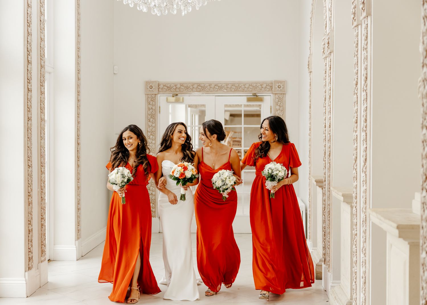 Bridesmaids in flowing red dresses pose together in an elegant white room with ornate moldings and chandeliers.