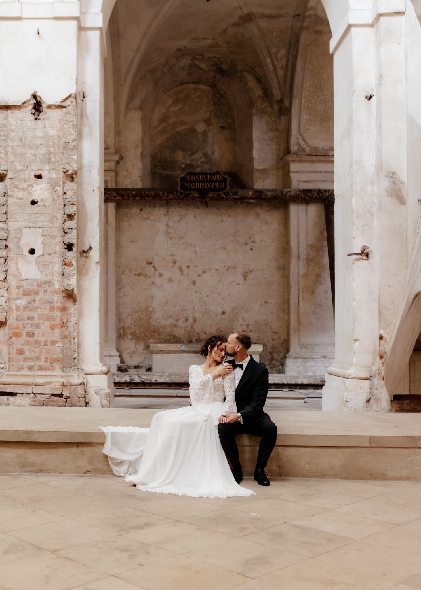 A couple in formal wedding attire shares a romantic moment in an old stone building with arched columns and brick walls.