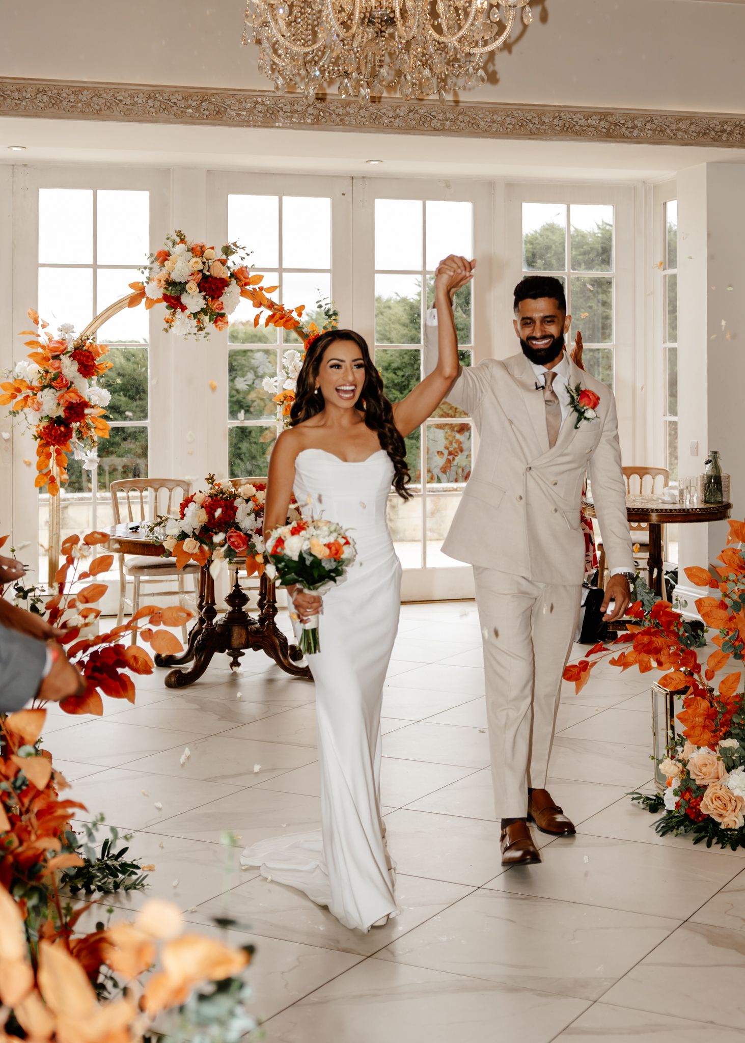 Wedding party dances in an elegant white ballroom decorated with orange and peach floral arrangements and chandeliers.