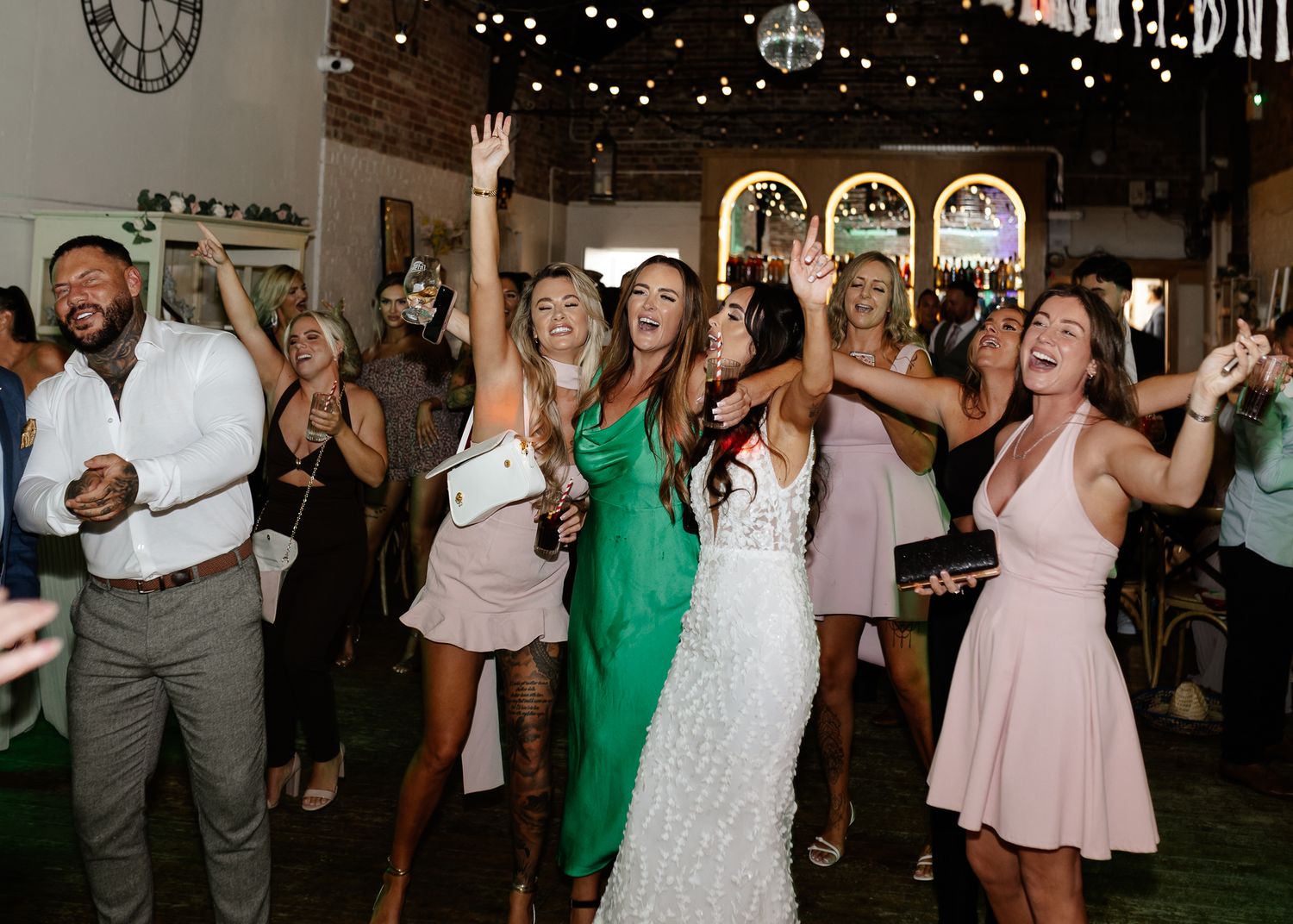 Wedding guests dance and celebrate with raised arms in a venue decorated with twinkling lights and arched windows.