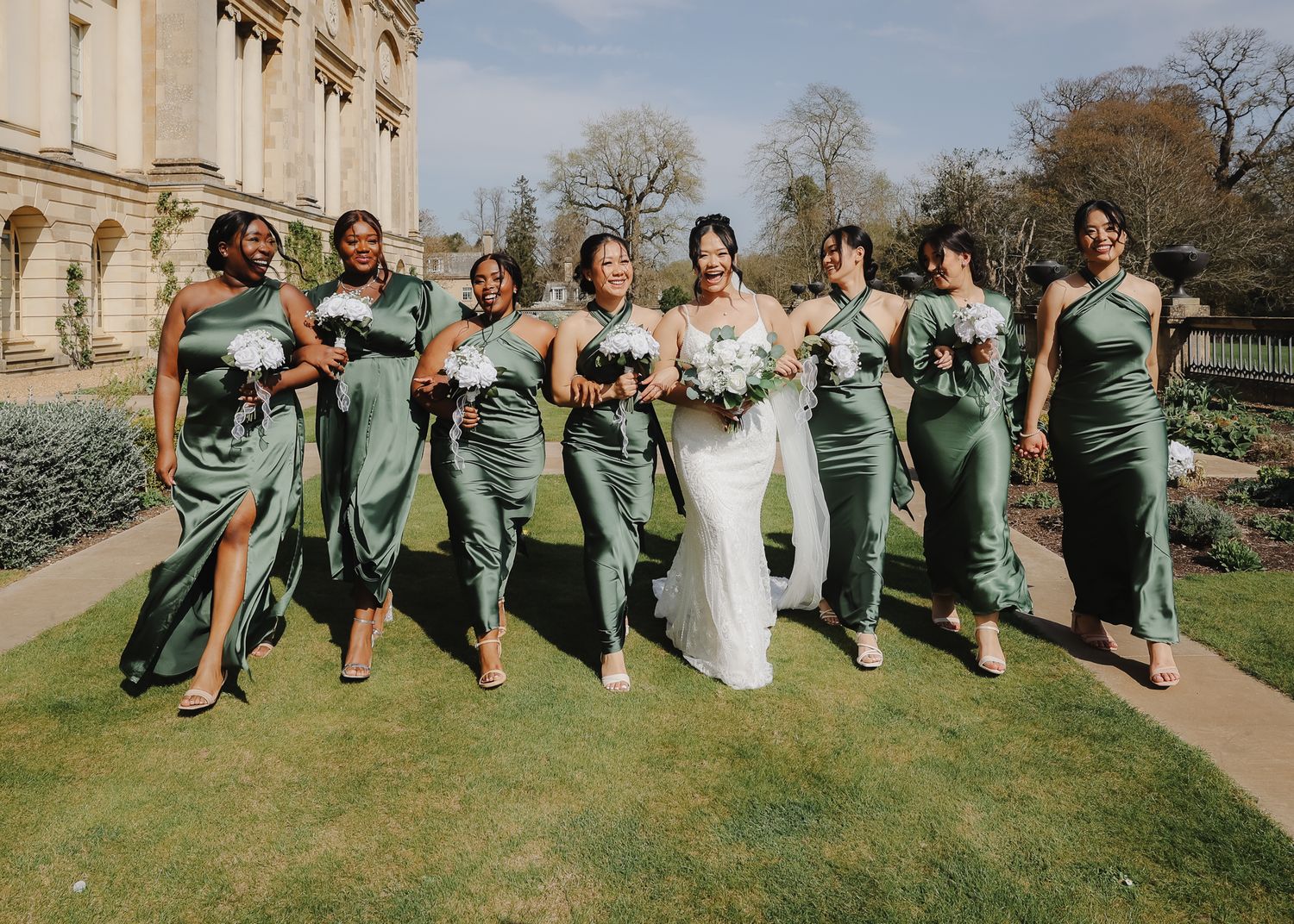 Bridal party in sage green dresses celebrates joyfully on sunny lawn outside historic building.