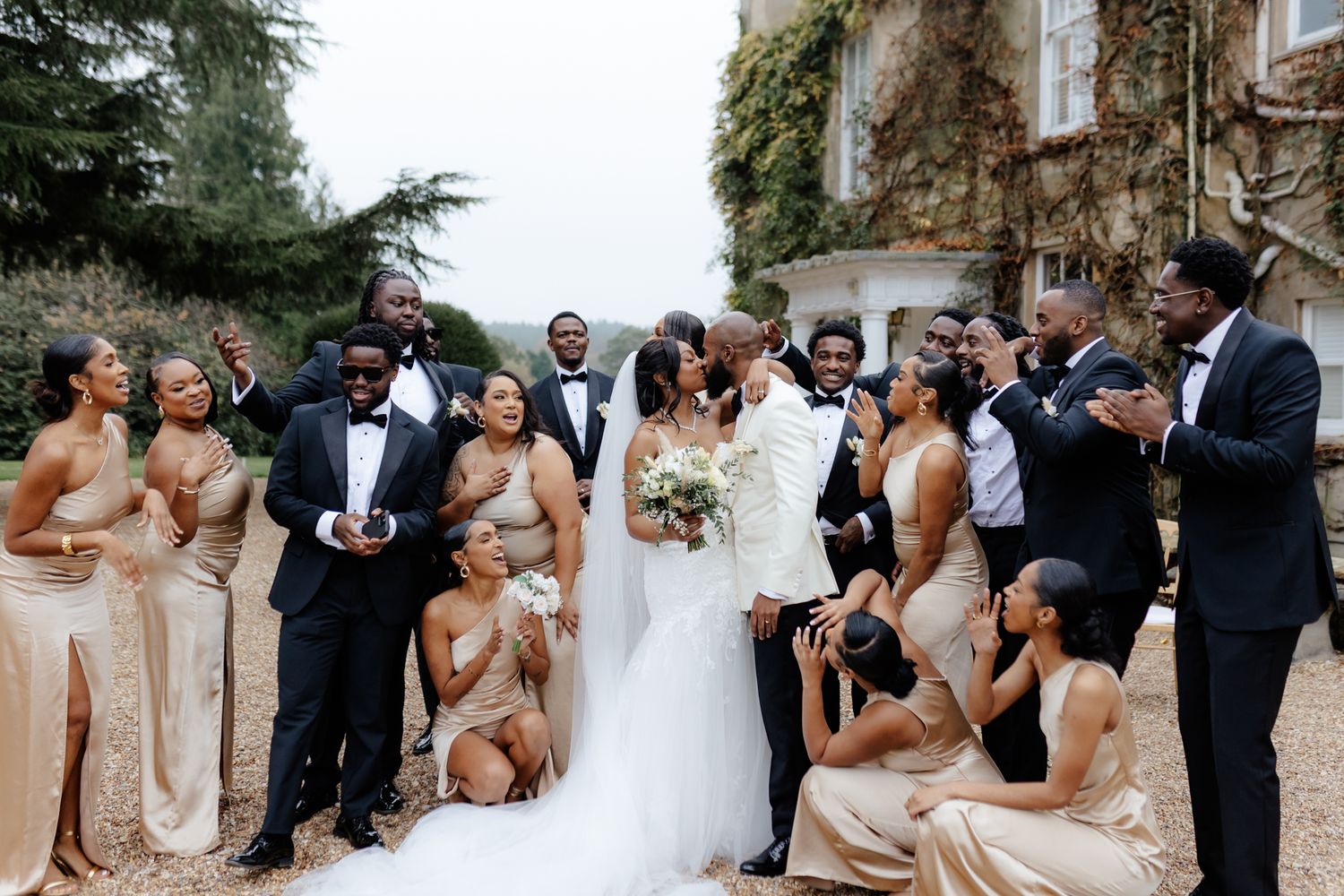 Wedding party members in blush pink dresses and black tuxedos pose together for celebratory photos outdoors.