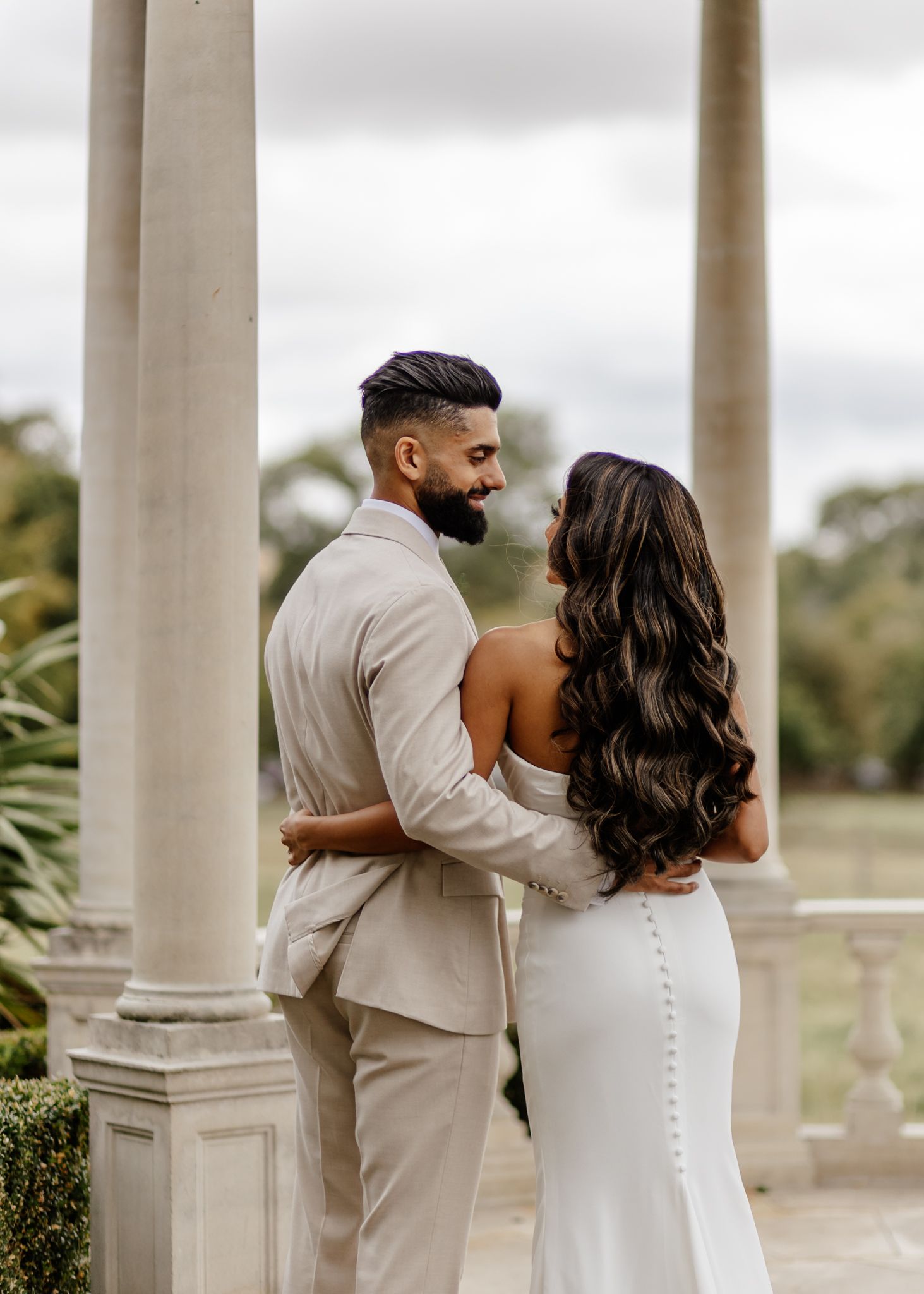 Elegant couple in beige and white wedding attire embrace between classic white columns on their special day.