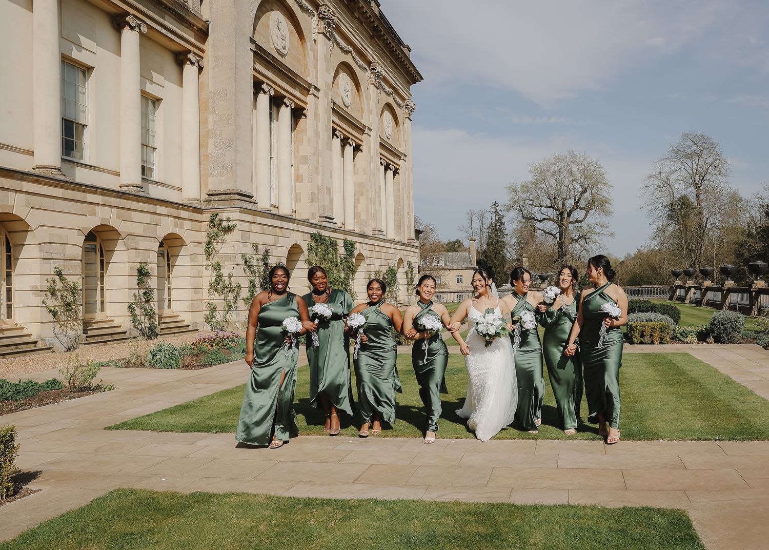 A wedding party in sage green dresses celebrates joyfully on the lawn of an elegant historic mansion on a sunny day.