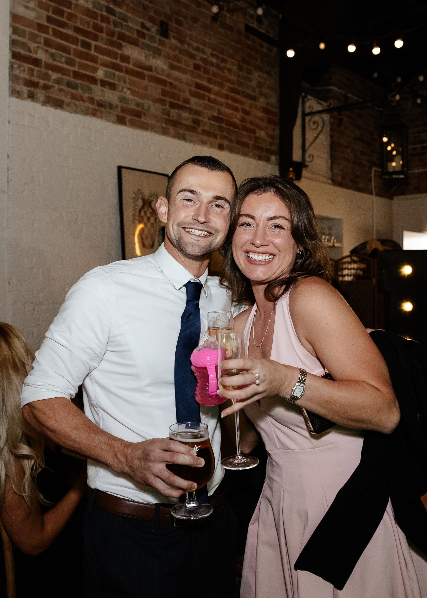 Two people in formal attire smile while holding cocktails at an indoor event with exposed brick walls and string lights.