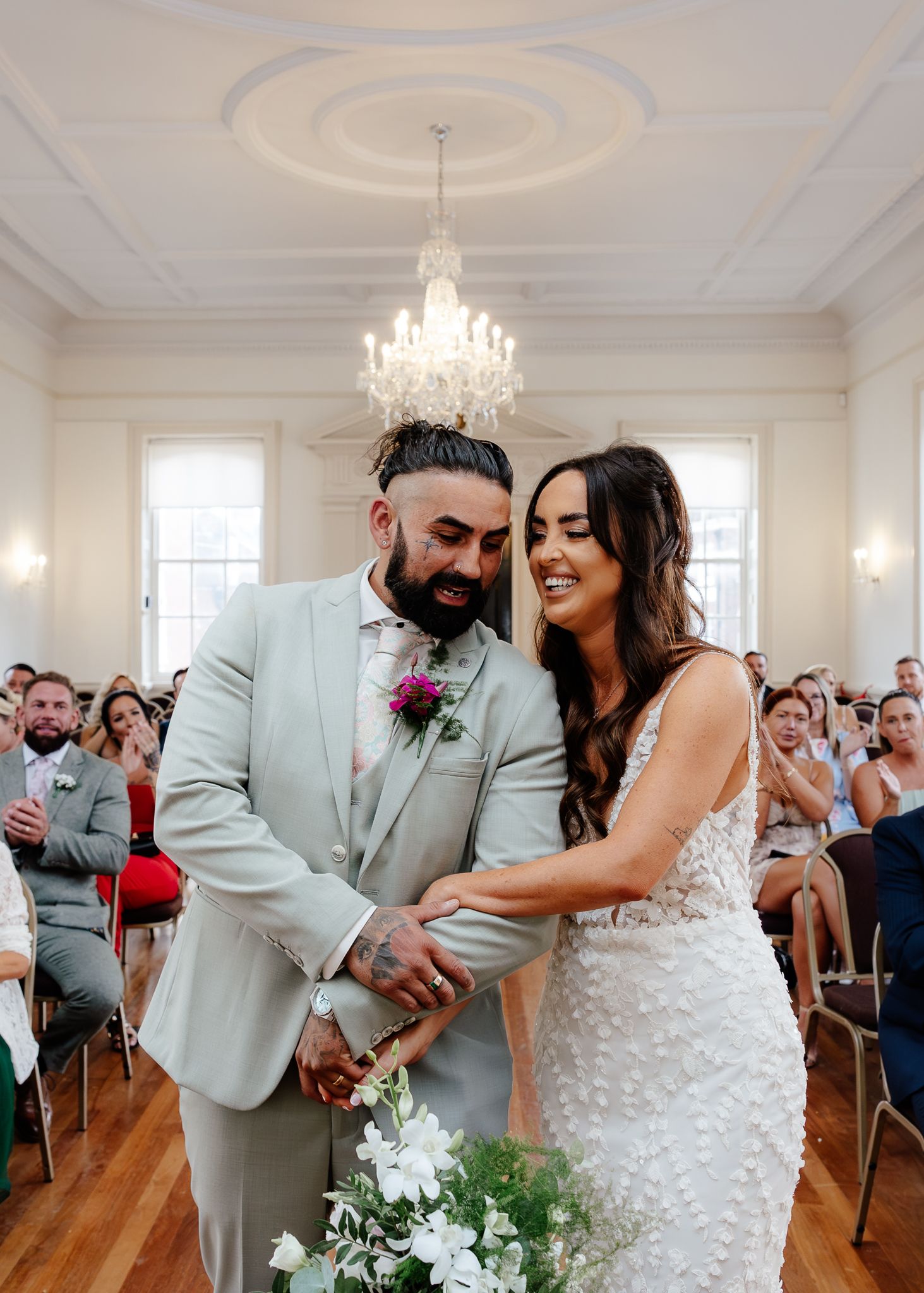 A couple shares a joyful moment during their wedding ceremony in an elegant room with a crystal chandelier.