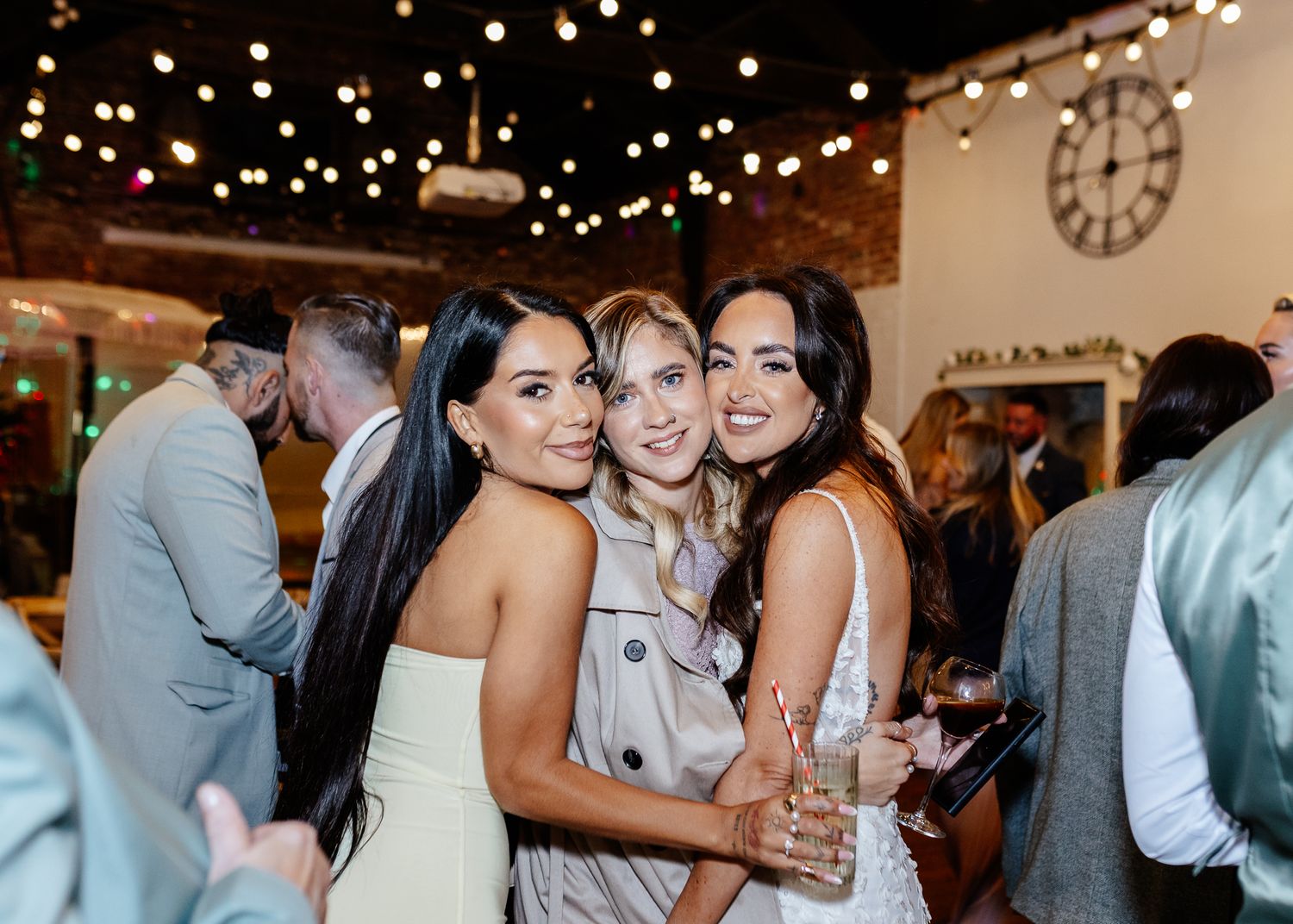 Three guests pose together during a festive indoor celebration with string lights and a clock on the wall.