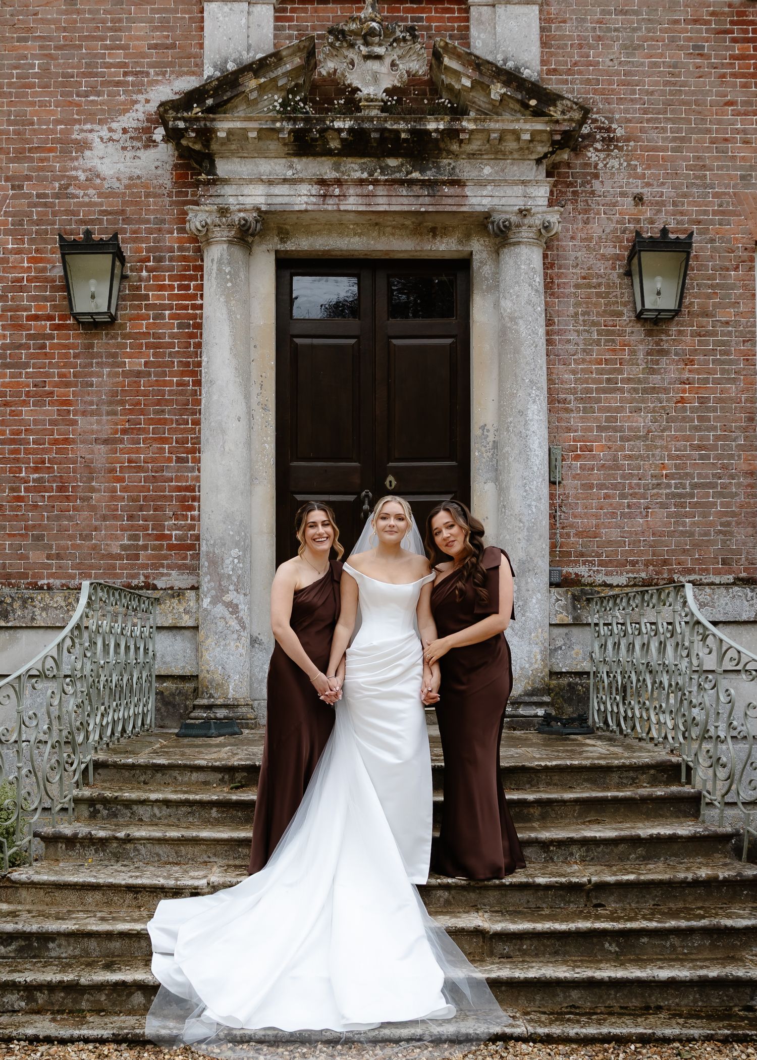 Bride in white gown with two bridesmaids in brown dresses on stone steps of elegant brick manor.