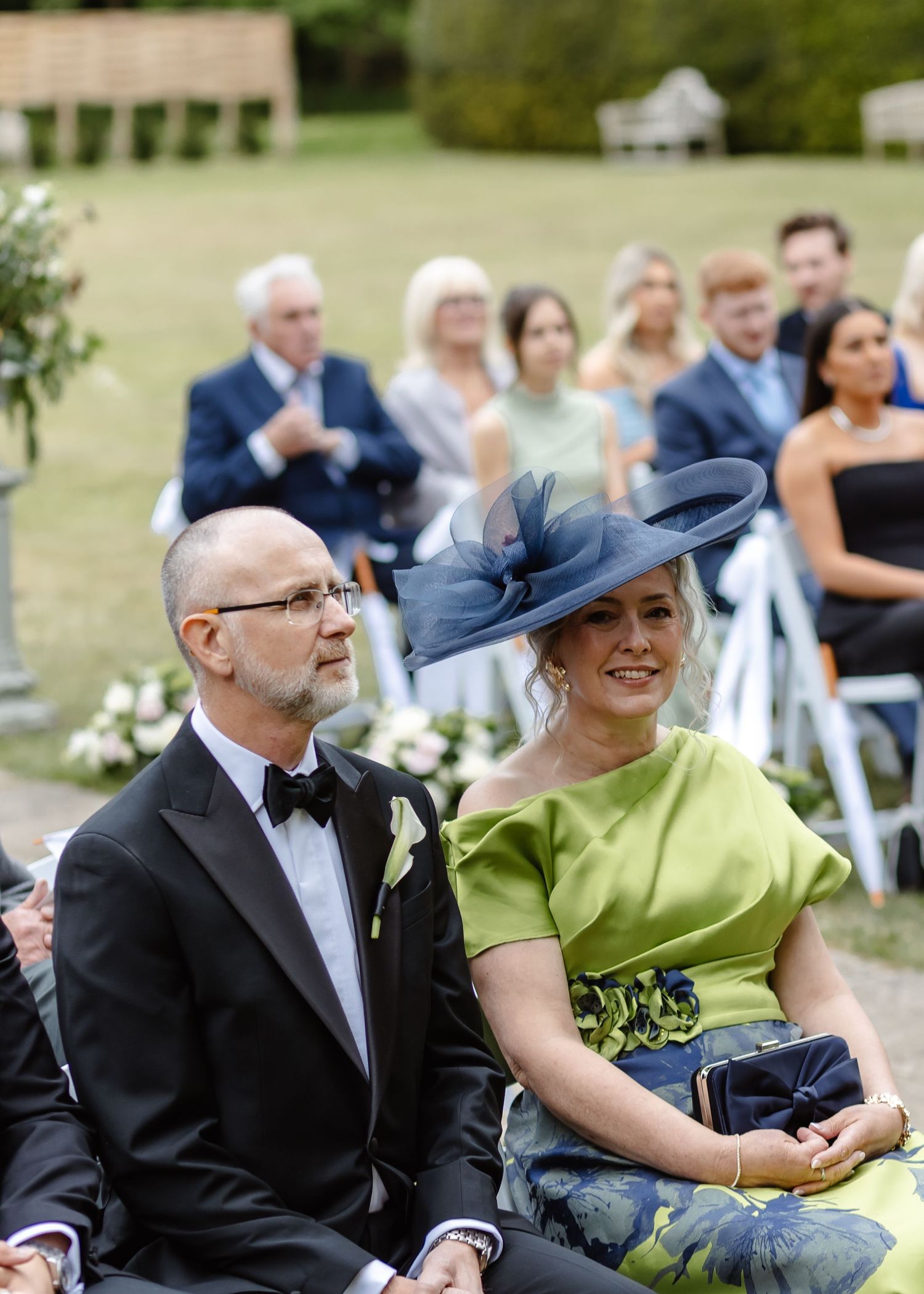 Elegant wedding guests seated outdoors, woman in green dress and navy fascinator hat smiling during ceremony.