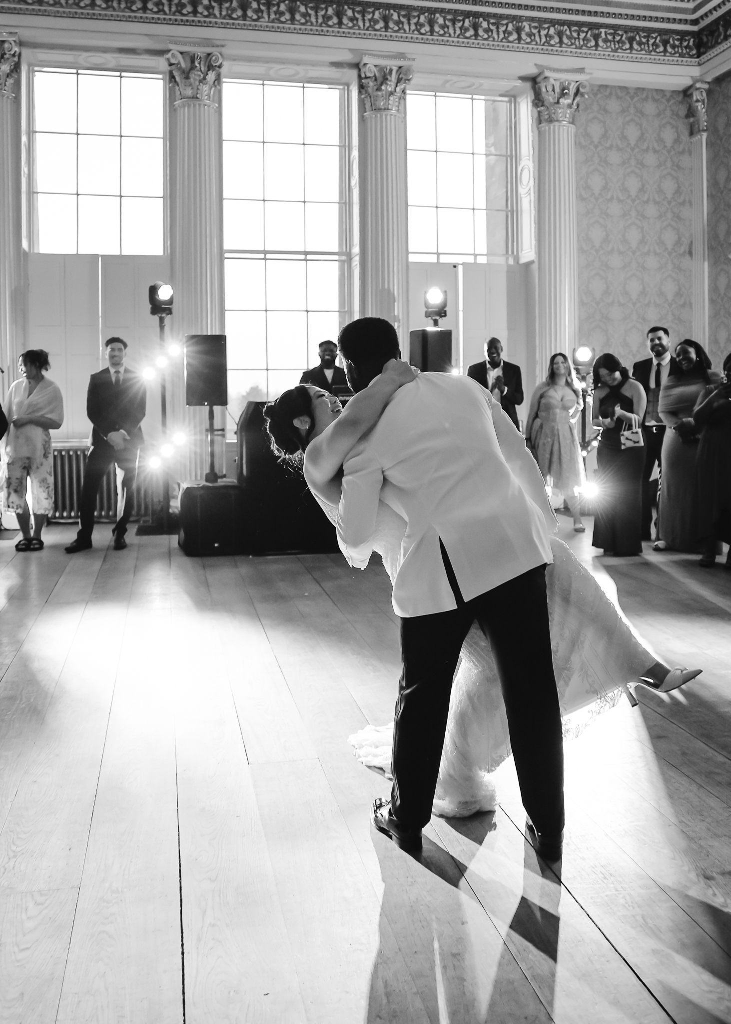 Black and white photo of couples dancing in a bright ballroom with tall windows.