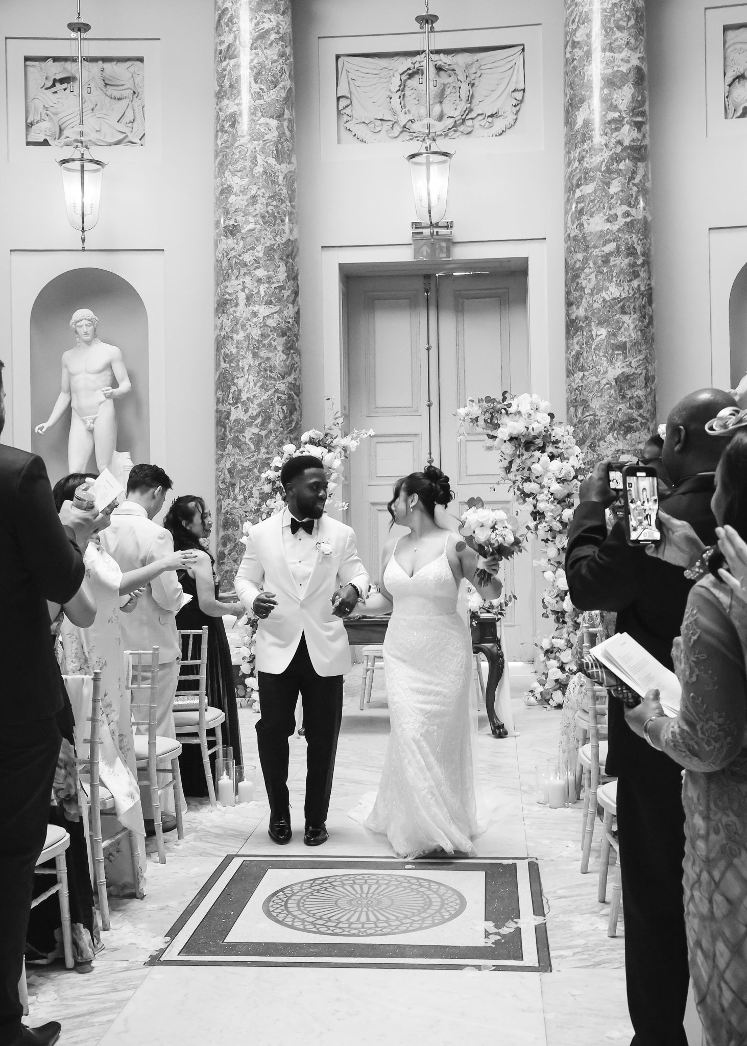 Wedding reception in a classical venue with marble pillars and guests gathered around the dance floor.