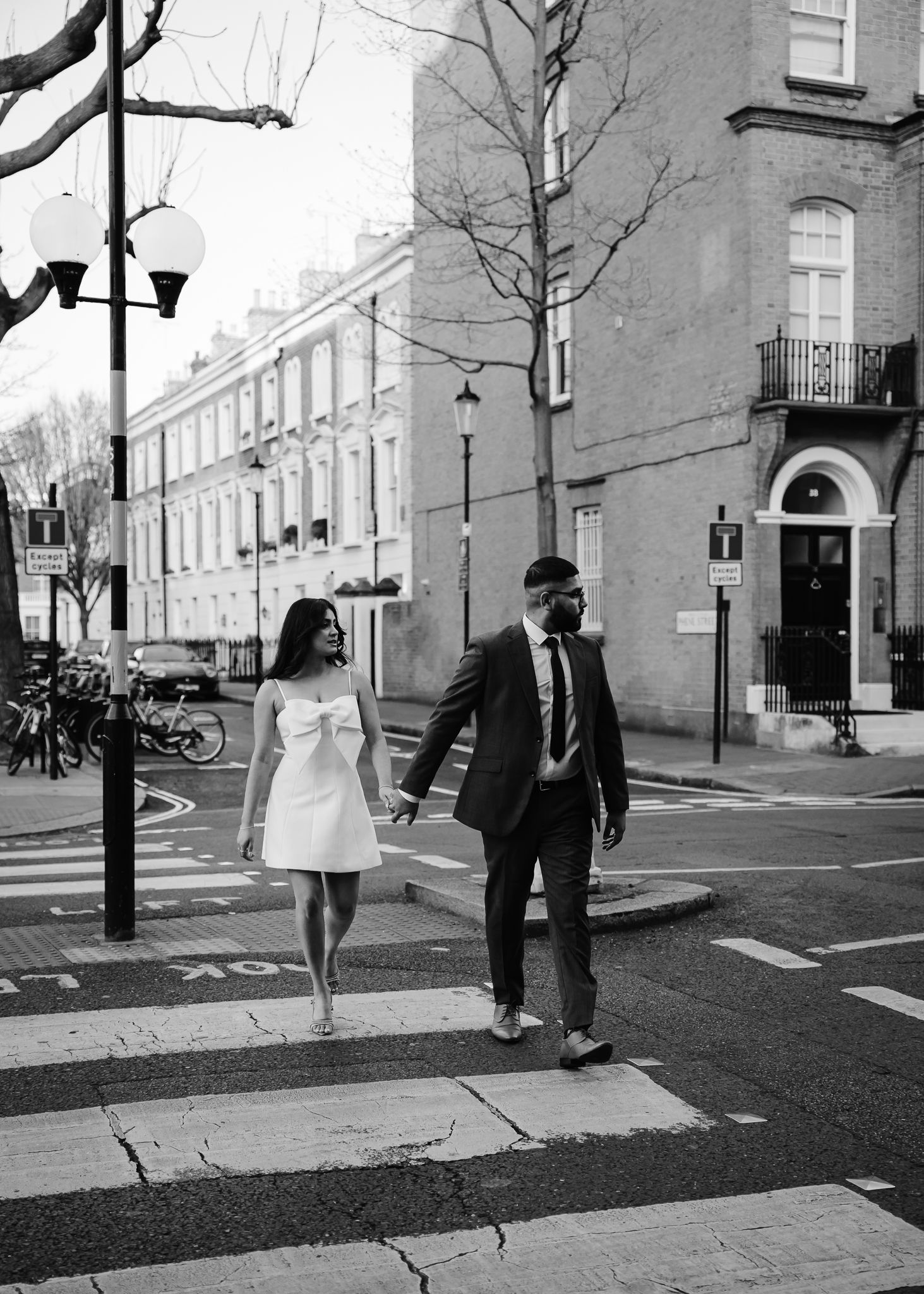 Black and white street scene showing people walking past brick buildings on an urban sidewalk with bare trees.