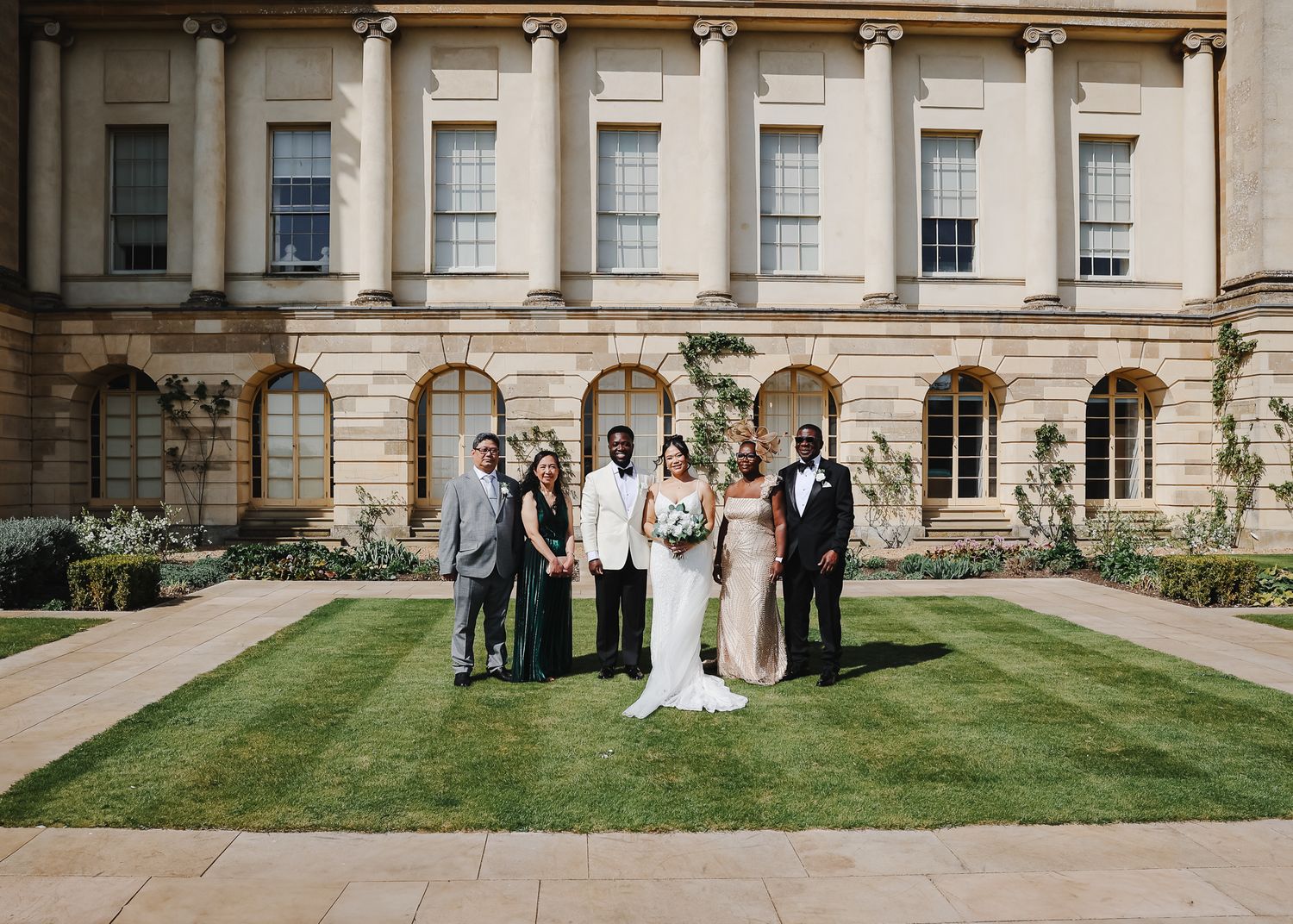 Wedding party stands on the manicured lawn in front of a grand stone mansion with arched windows and columns.