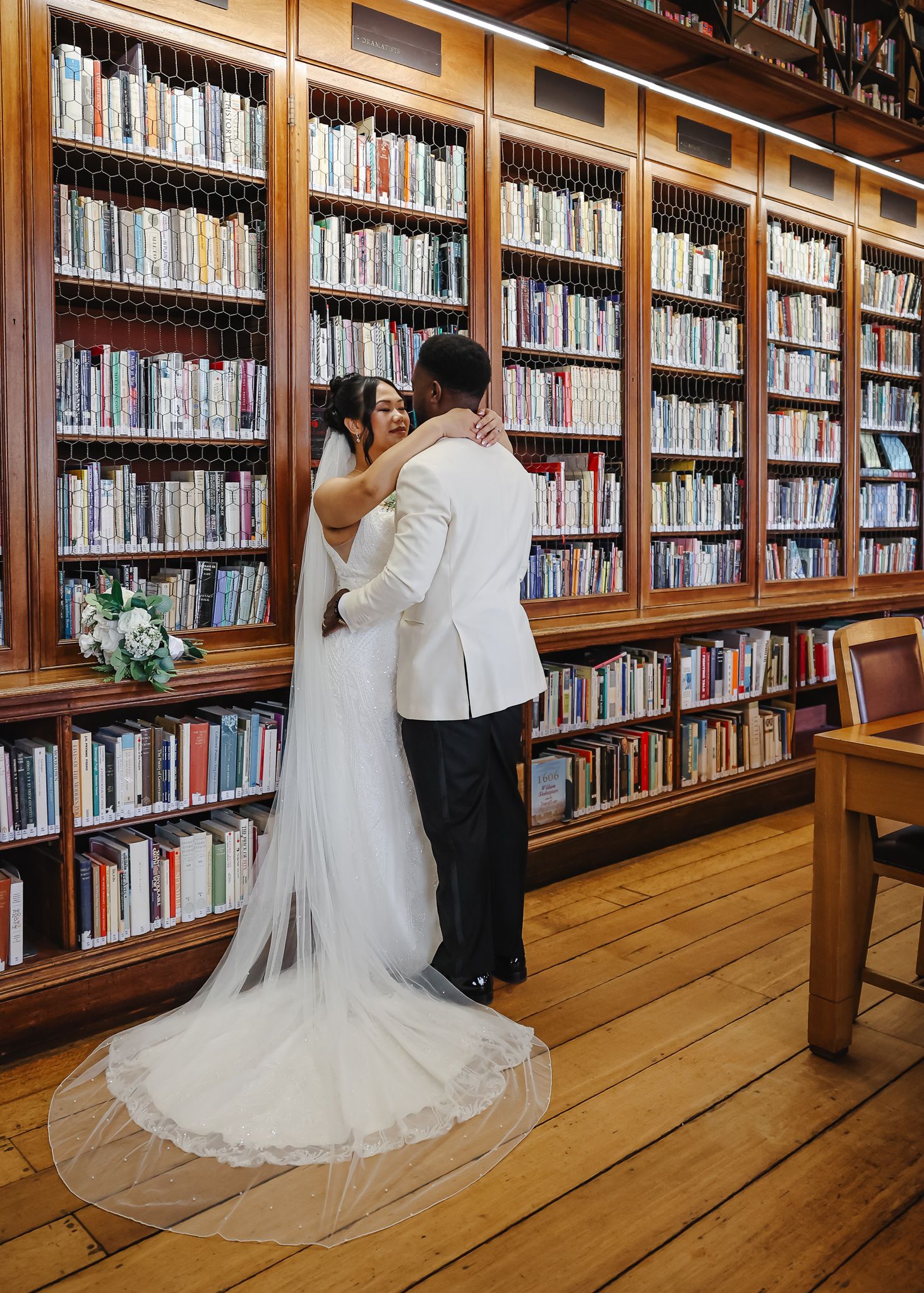A couple in wedding attire browses books together in a wooden library with tall bookshelves and hardwood floors.