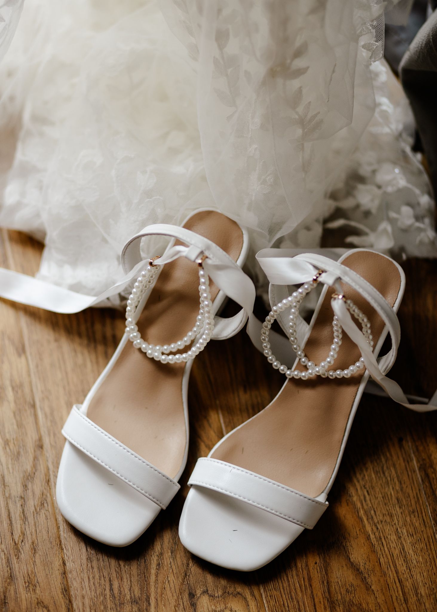 Elegant white bridal sandals with pearl straps displayed on wooden floor beneath flowing wedding dress fabric.