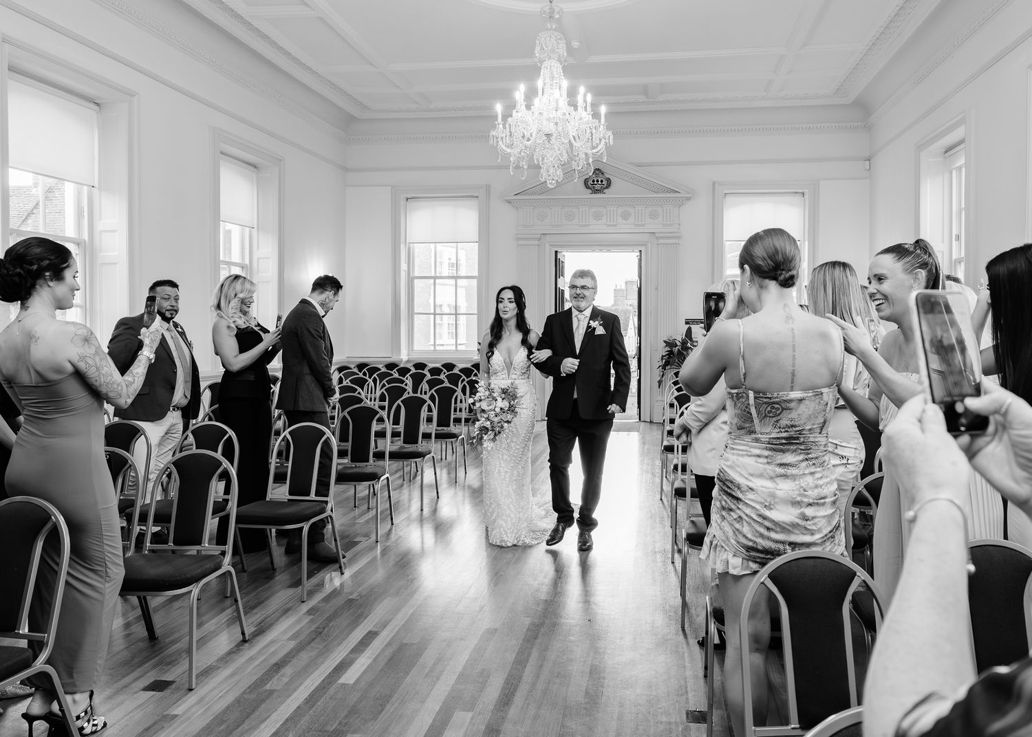 Wedding guests stand and mingle in an elegant white ballroom with a crystal chandelier and hardwood floors.