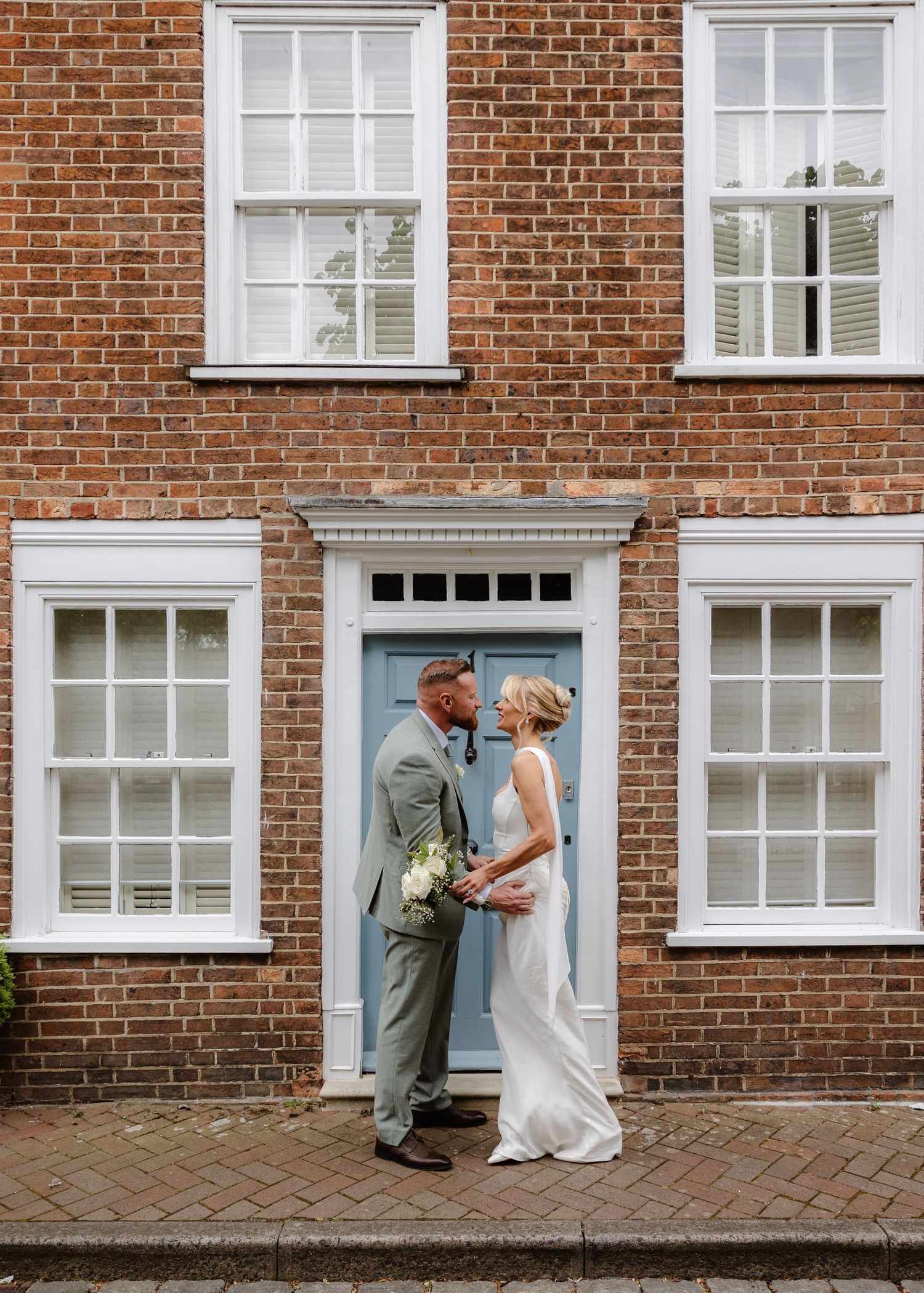 Bride and groom stand facing each other holding bouquet before blue door of red brick building on cobblestone street.