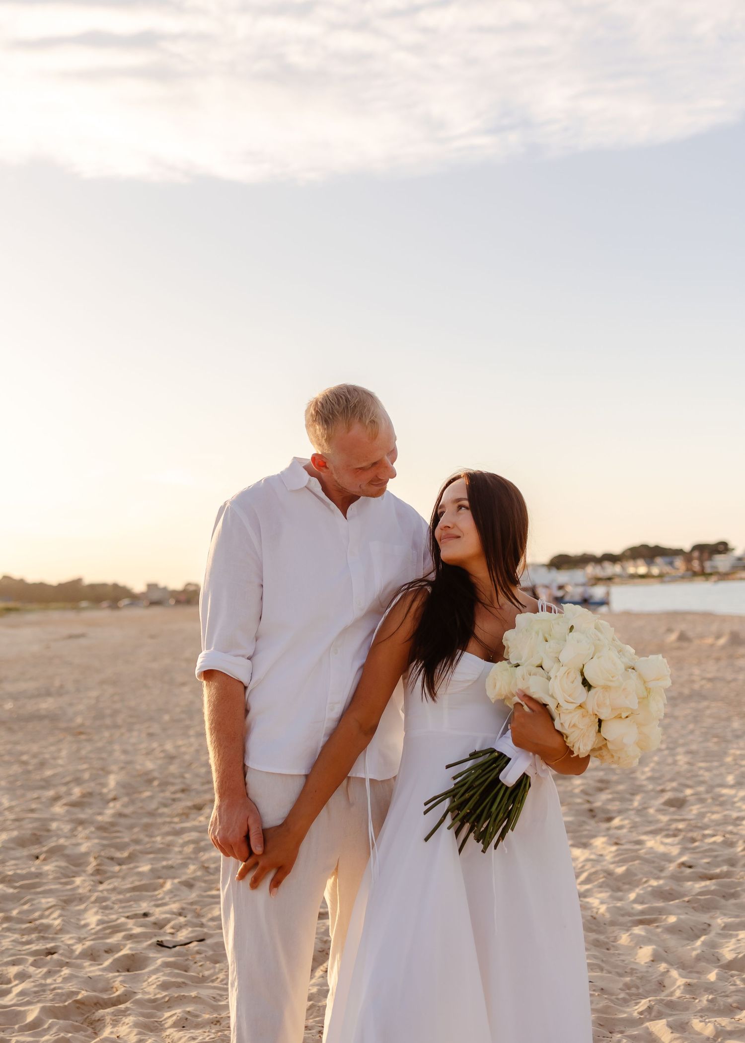 Couple in white outfits on beach at sunset, bride holding white roses bouquet, romantic seaside wedding photo.