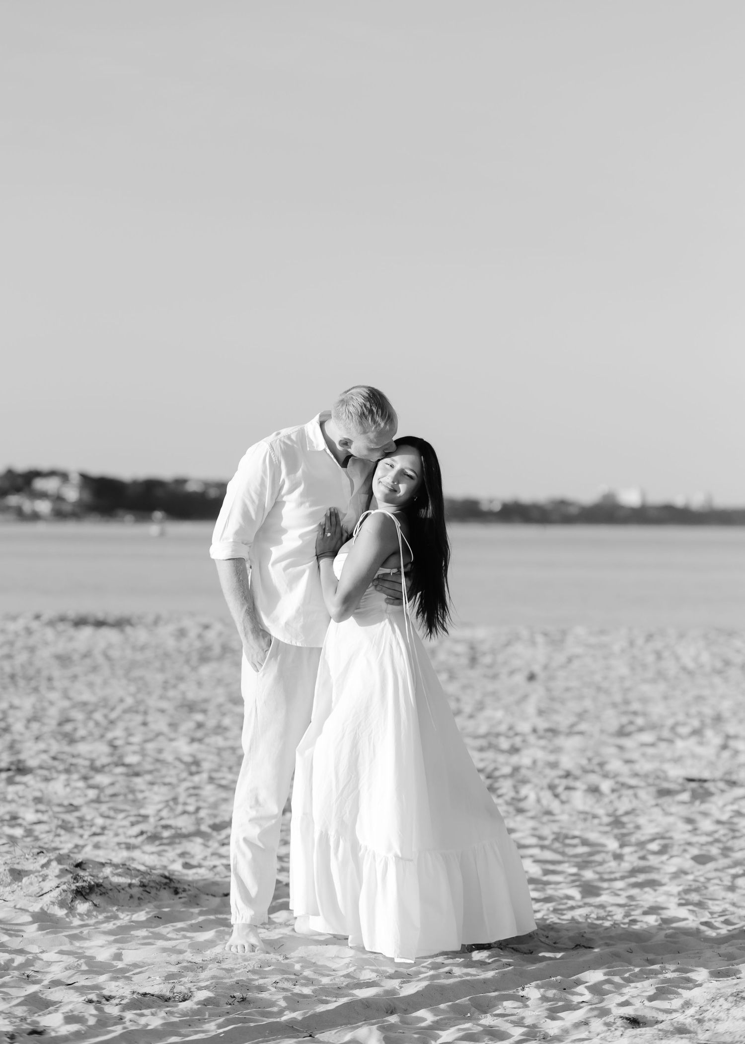 Black and white photo of a couple embracing on a sandy beach, man in white shirt kissing woman in flowing white dress.