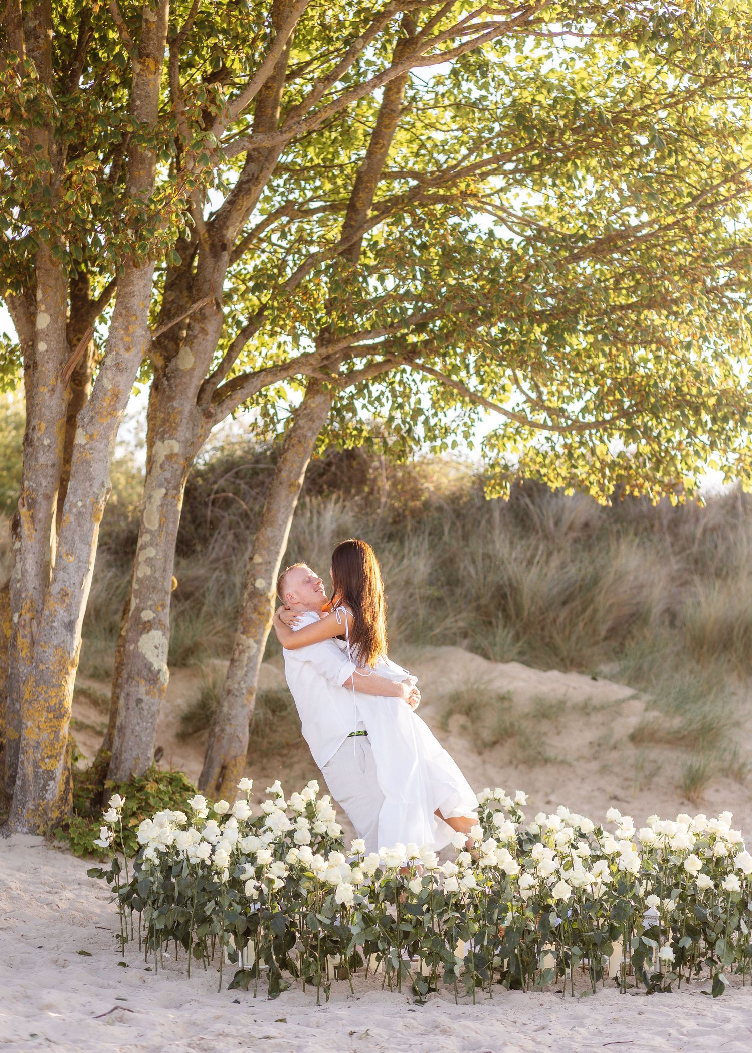 Couple embracing among white flowers on a sandy beach at golden hour, surrounded by trees.