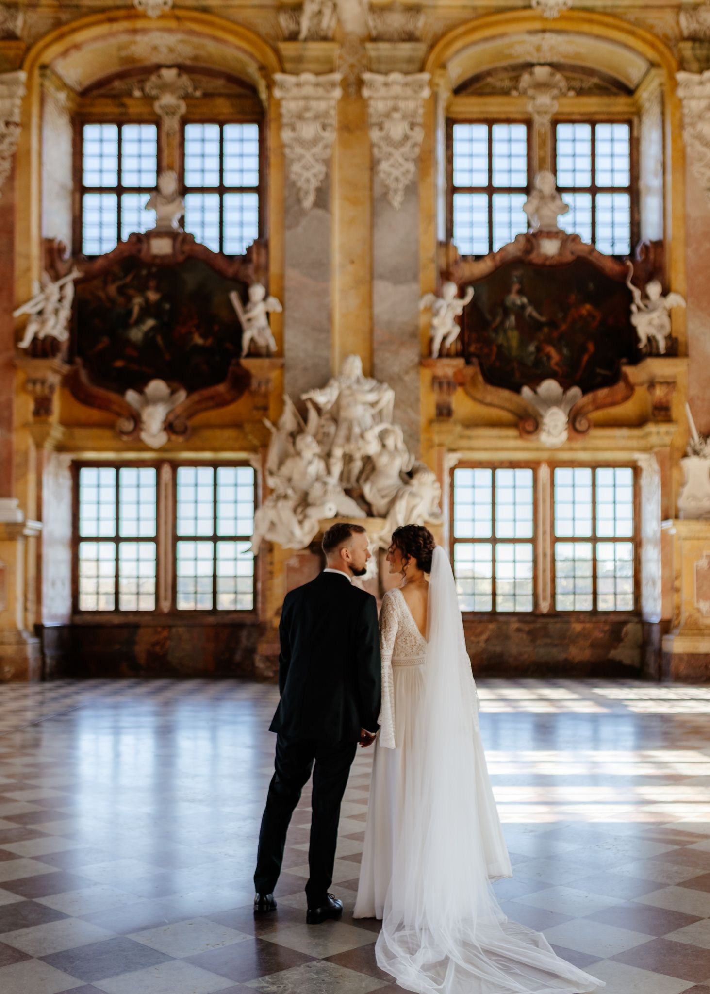 A couple dances in an elegant ballroom with ornate architecture, tall windows, and natural light streaming in.