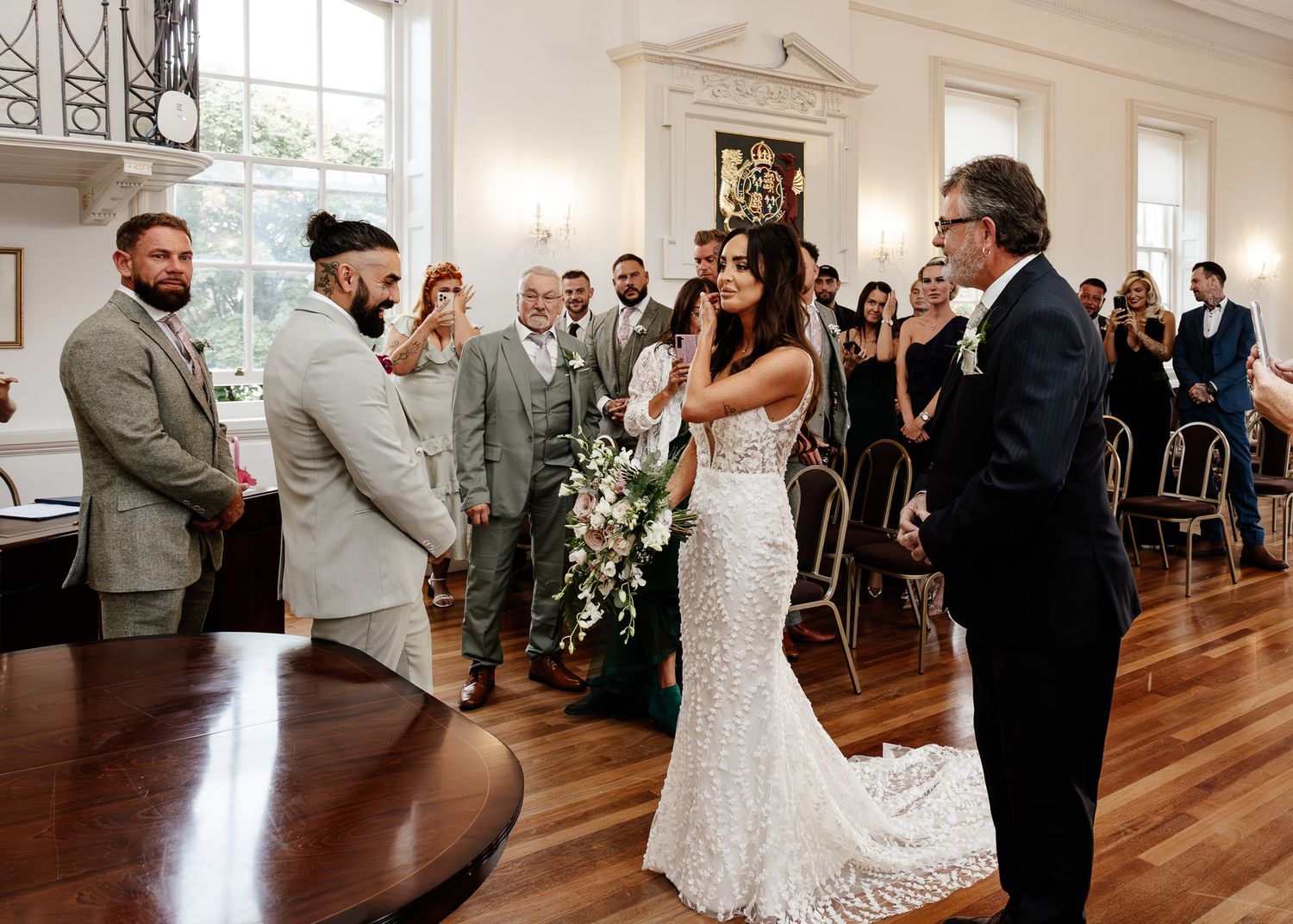 A wedding ceremony takes place in a bright, classic room with hardwood floors and large windows.