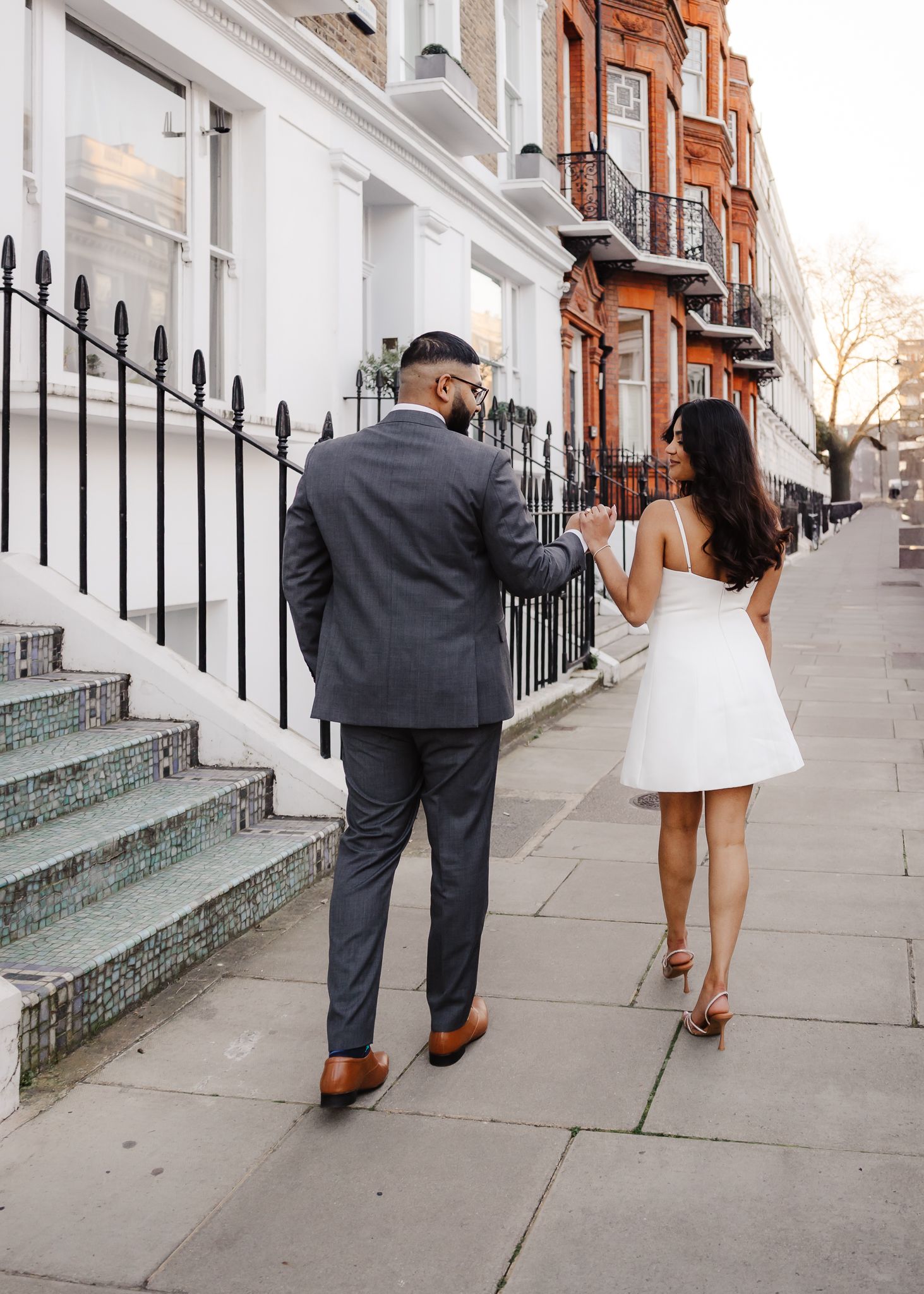 A couple walking down a London street with classic white townhouses and red brick buildings in the background.