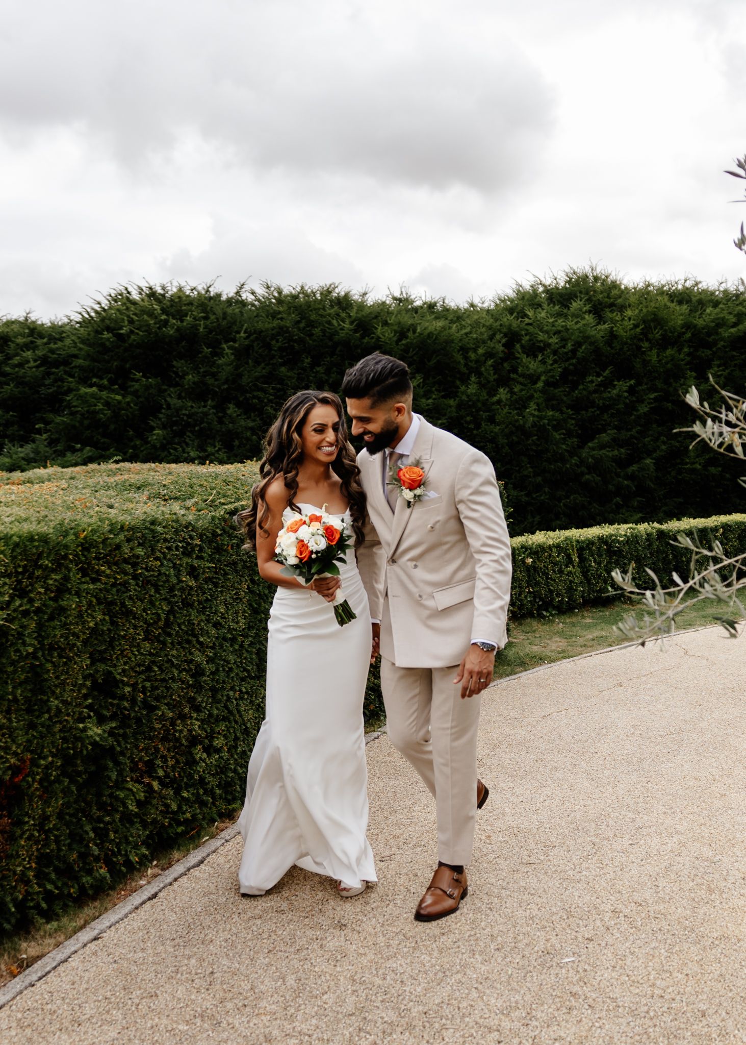A couple in wedding attire shares a romantic moment on a gravel path bordered by manicured hedges.