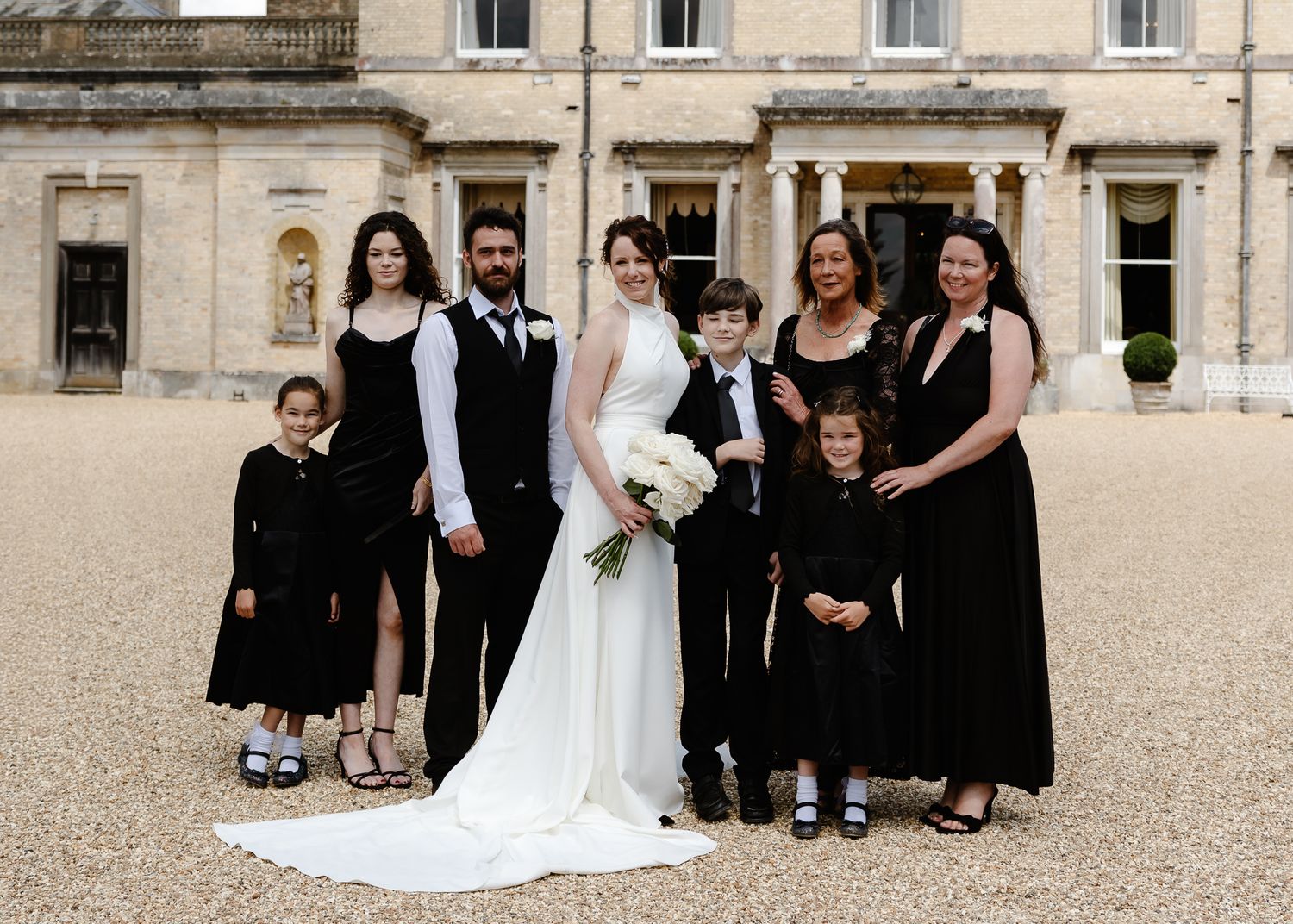 Wedding party in formal attire poses together outside a grand stone building.