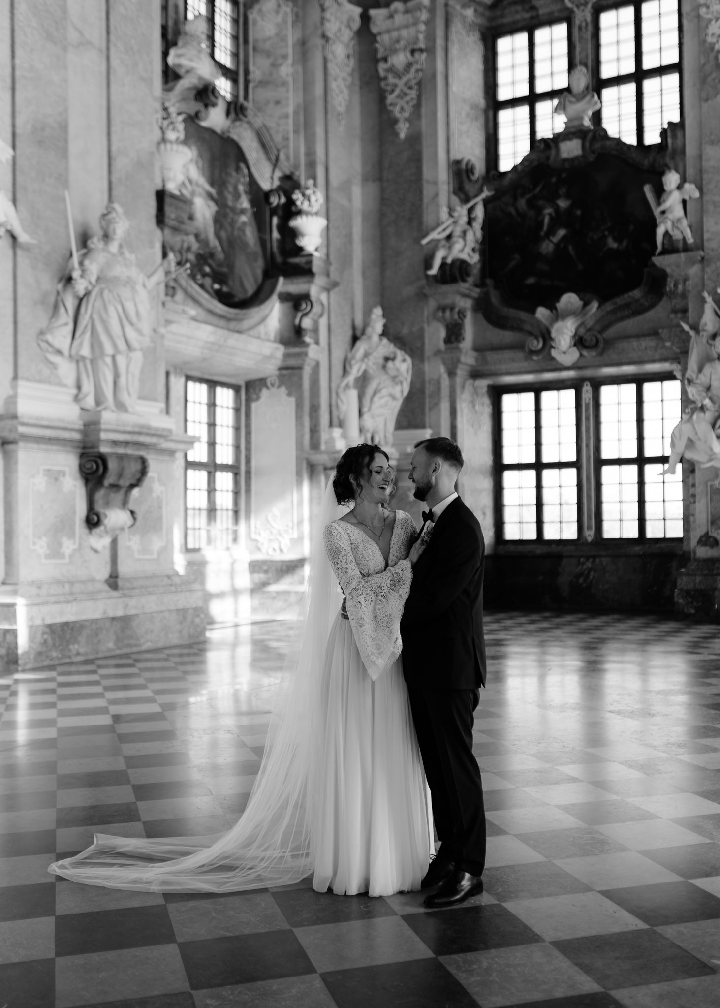 A couple in wedding attire dancing in an elegant baroque palace hall with ornate white sculptures and tall windows.