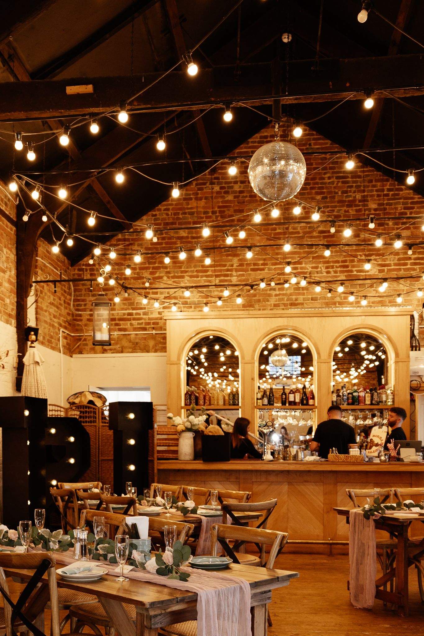 Wedding reception venue with exposed brick walls, string lights, and elegant table settings in a rustic industrial space.