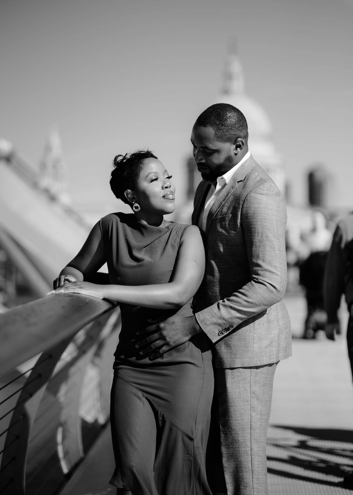 A romantic black and white photo of a couple laughing together on a sunny day with architectural details in background.