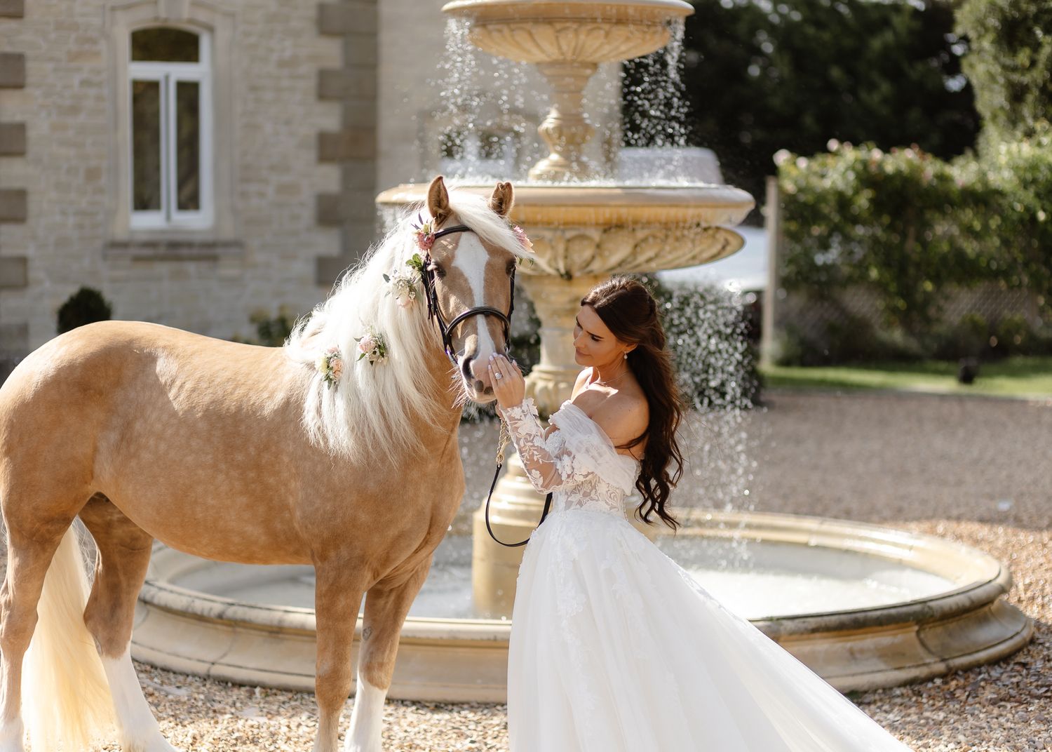 Bride in white gown feeding a palomino horse by an ornate stone fountain at an elegant chateau wedding.
