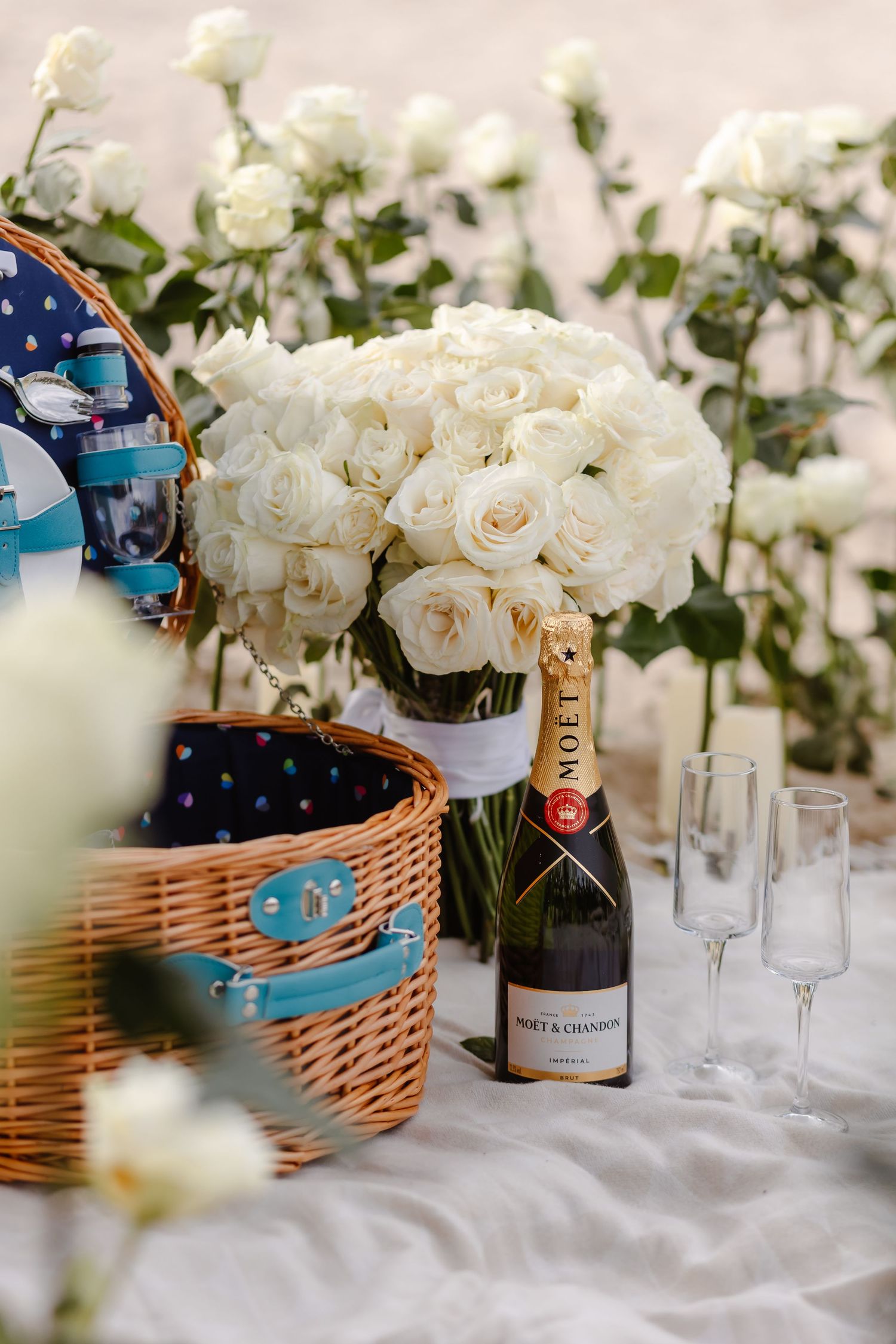 Elegant picnic setup with Moet Chandon champagne, crystal flutes, white roses bouquet, and wicker basket on white linen.